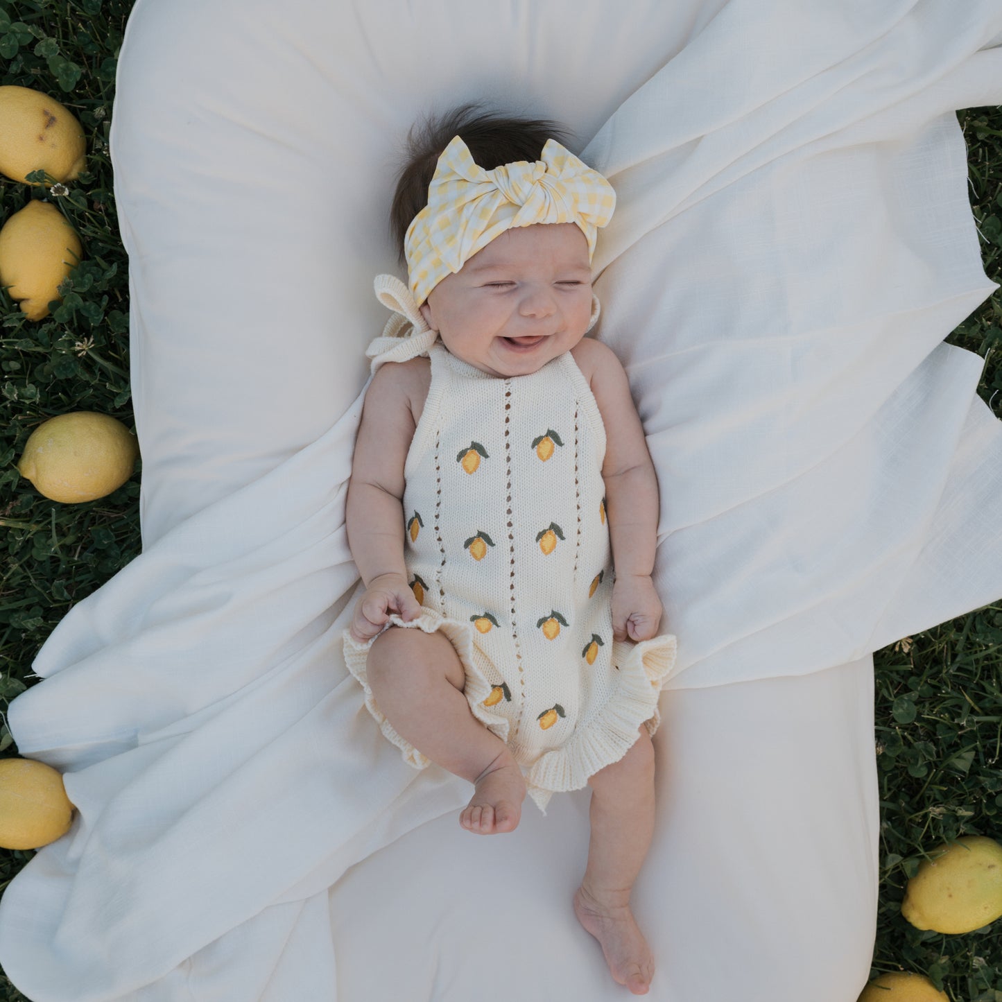 A baby in the forever french baby Knit Ruffle Romper | Lemon with adjustable tie straps and a yellow headband smiles with eyes closed, lying on a white blanket outdoors among lemons on green grass.