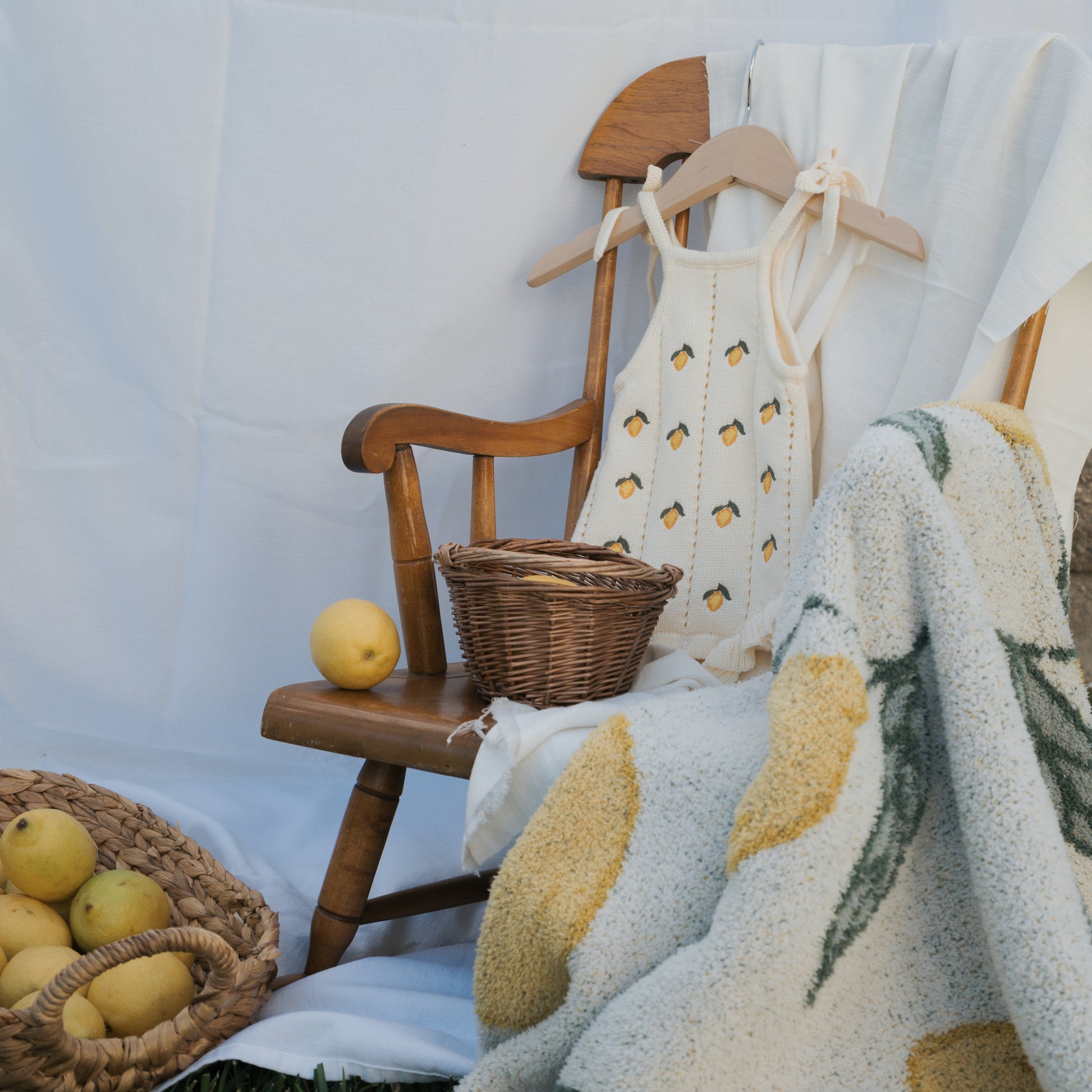 A small wooden chair showcases the forever french baby Knit Ruffle Romper | Lemon with adjustable tie straps and snaps, surrounded by yellow apples, a wicker basket, and a white-yellow blanket, all set against a white background.