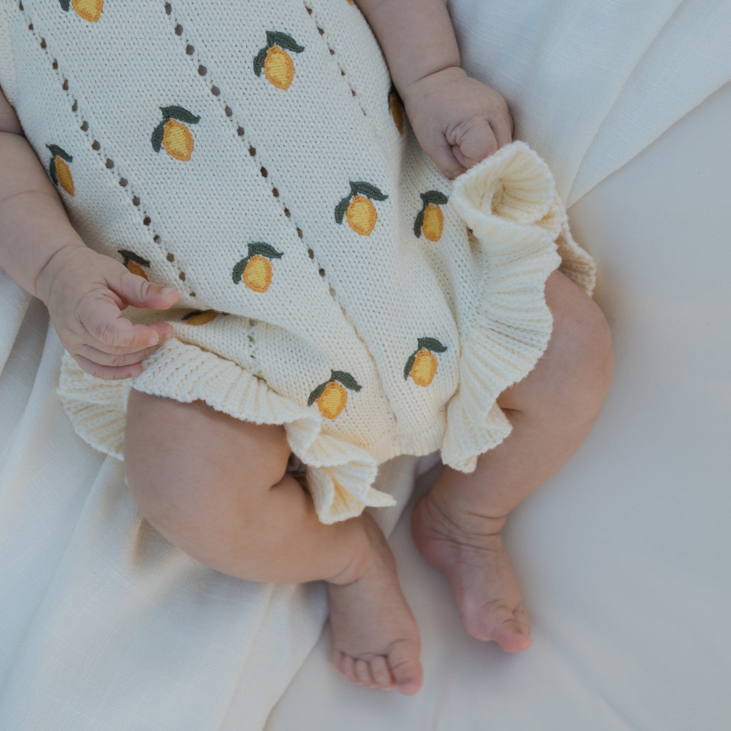 A baby wears the forever french baby Knit Ruffle Romper | Lemon, a cream-colored knit romper with lemon print, ruffled edges, and adjustable tie straps, while lying on soft white fabric with visible hands and legs.