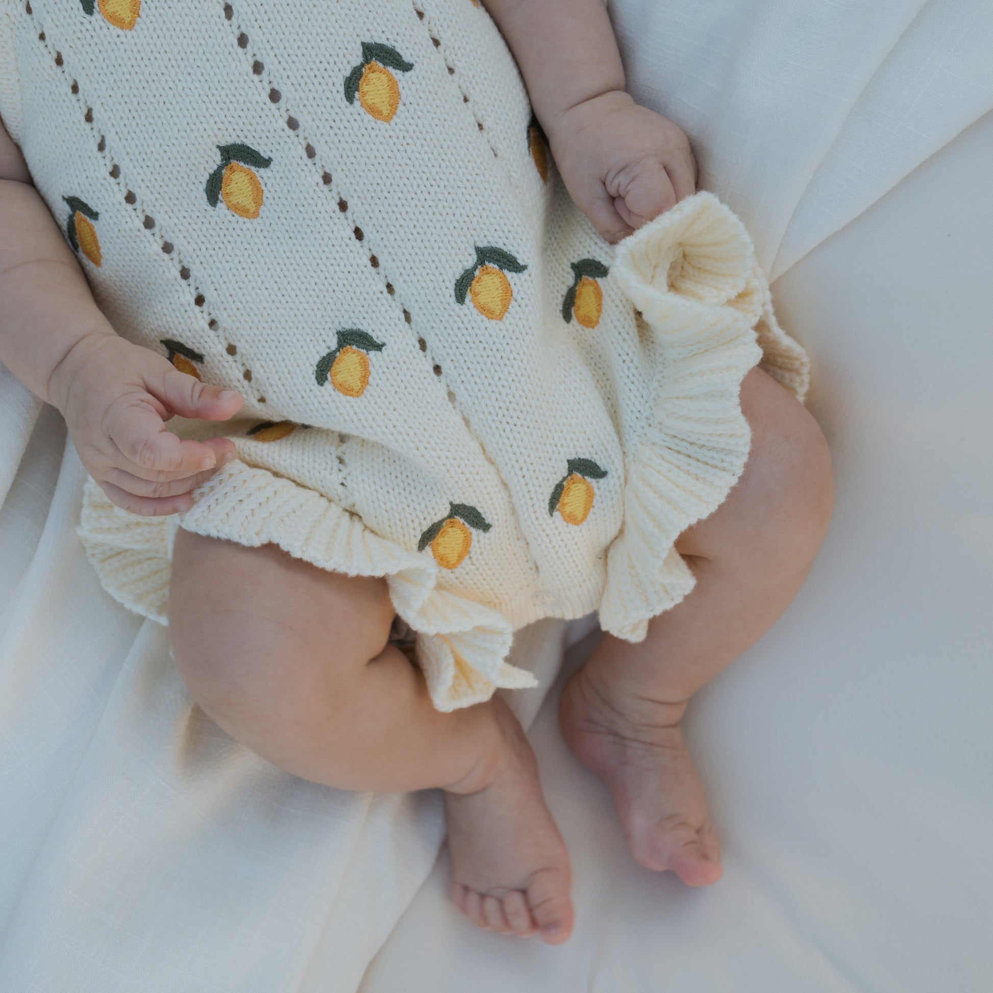 A baby wears the forever french baby Knit Ruffle Romper | Lemon, a cream-colored knit romper with lemon print, ruffled edges, and adjustable tie straps, while lying on soft white fabric with visible hands and legs.