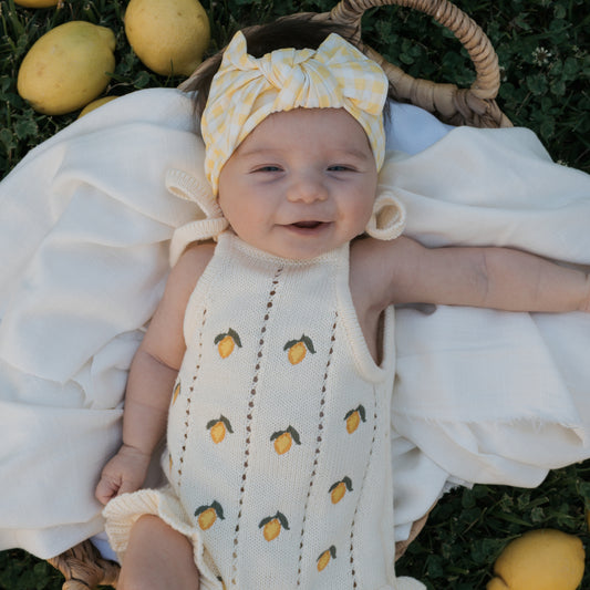 A smiling baby lies on a white blanket in a basket, wearing the forever french baby Knit Ruffle Romper | Lemon with adjustable tie straps and a yellow gingham headband. Lemons are scattered on the grass.