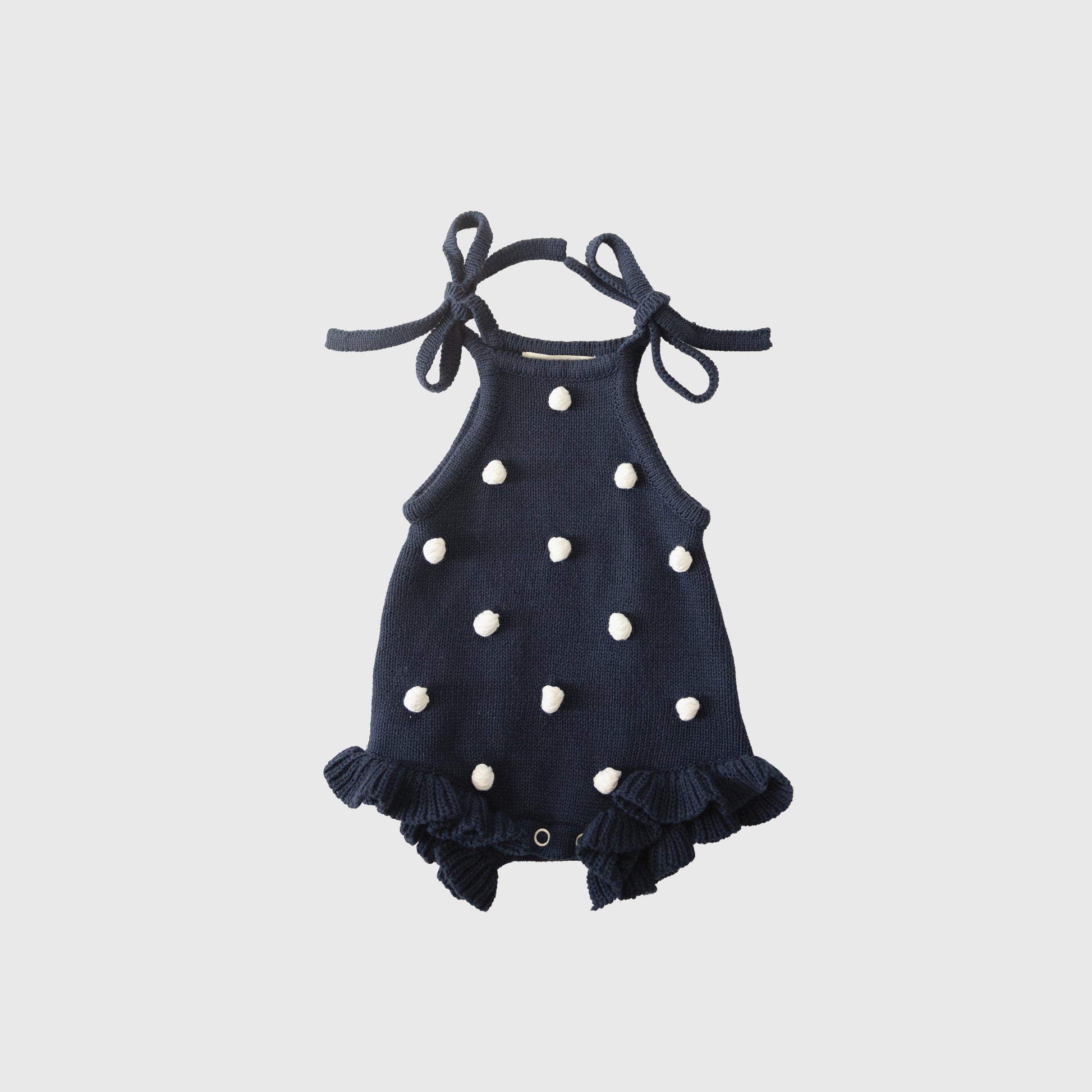 The forever french baby Knit Ruffle Romper | Emery, in navy blue knit with white pom-poms, ruffled legs, adjustable tie straps, and snap closures, is shown laid flat on a light gray background.