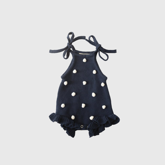 The forever french baby Knit Ruffle Romper | Emery, in navy blue knit with white pom-poms, ruffled legs, adjustable tie straps, and snap closures, is shown laid flat on a light gray background.