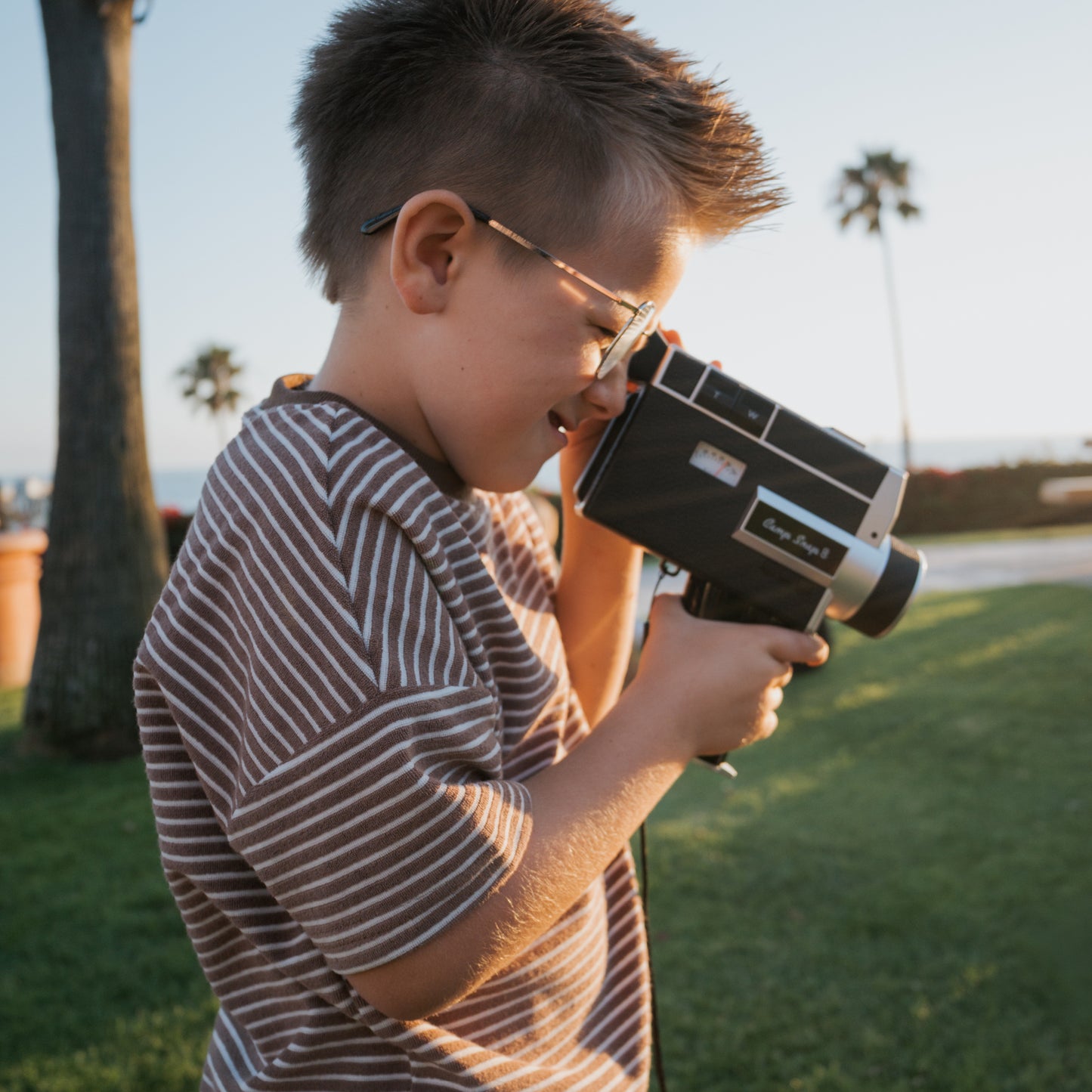A young boy in glasses wears the forever french baby Child Terry Short Set in Cola Stripe, filming outdoors with a vintage video camera amid sunny palm trees and grass.