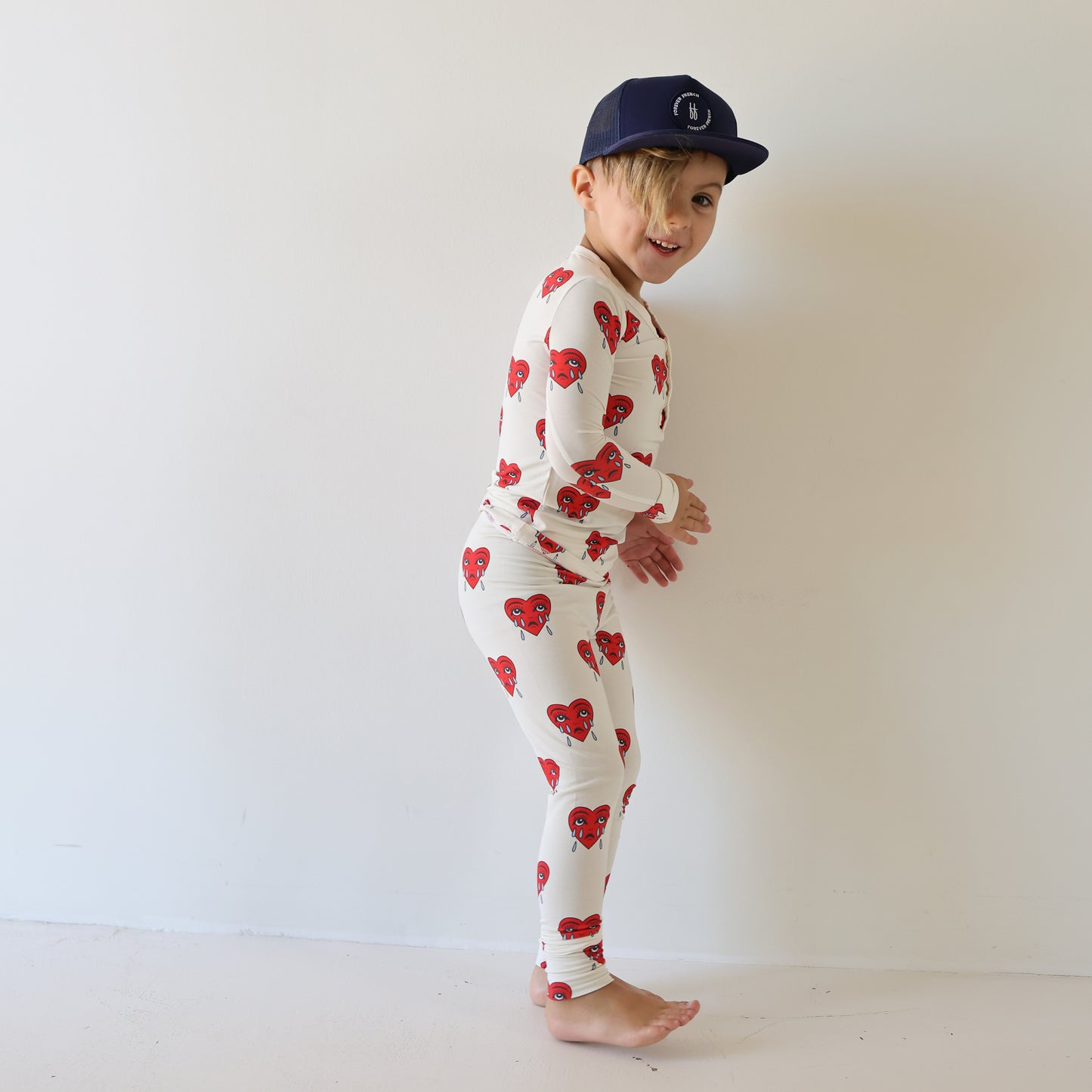 A young child in forever french baby's Bamboo Two Piece Pajamas | Sad Girl—white with red hearts—smiles barefoot in front of a plain white wall, wearing a dark blue baseball cap and looking over their shoulder.