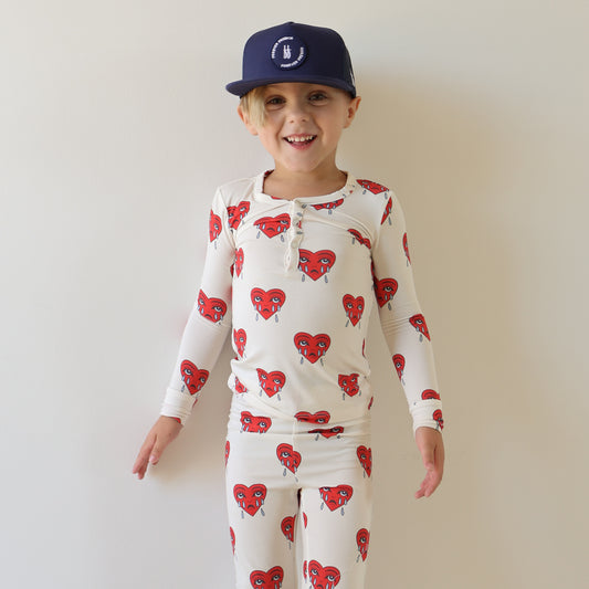 A young child smiles in front of a white wall, wearing forever french baby’s Bamboo Two Piece Pajamas | Sad Girl—navy blue cap and soft white bamboo sleepwear with red heart-shaped faces, perfect breathable pajamas for kids.