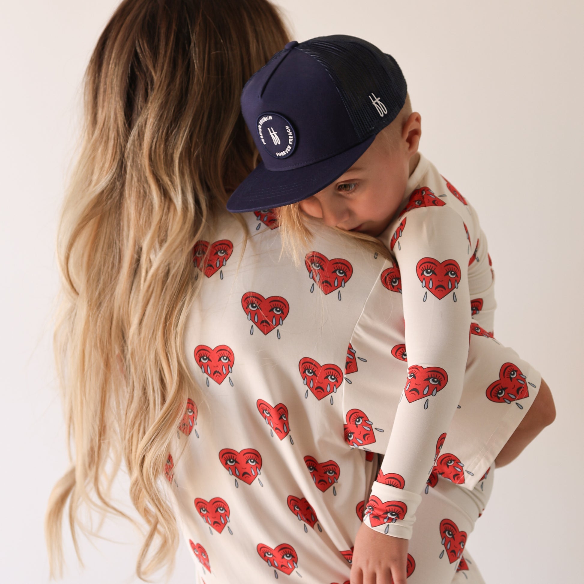 A woman with long, wavy blonde hair holds a child in a navy cap. Both wear forever french baby’s Bamboo Two Piece Pajamas | Sad Girl—cream, hypo-allergenic PJs with red crying heart designs—against a plain light background.