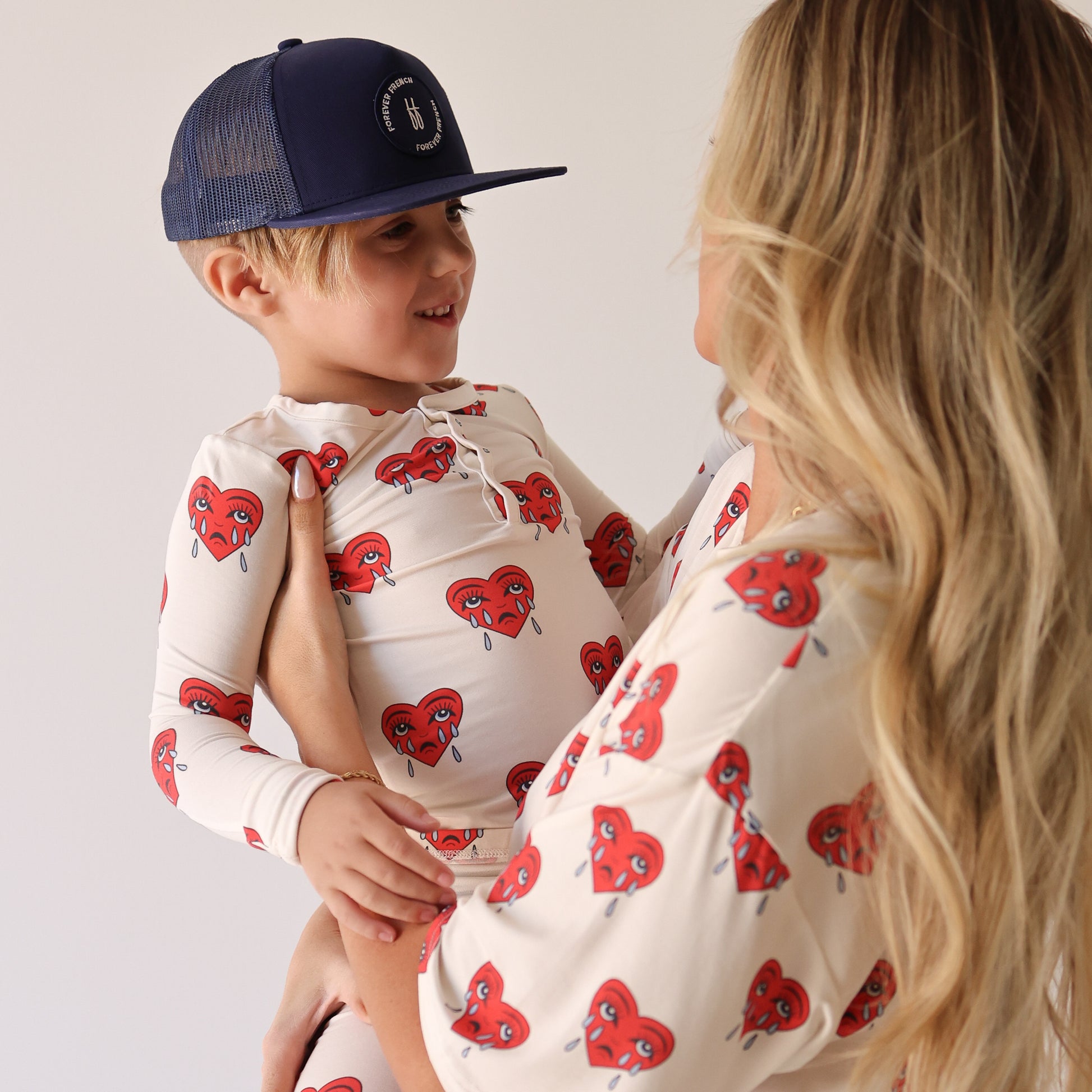 A blonde woman and a young child in navy cap smile at each other in matching forever french baby Sad Girl Short Sleeve Women's Bamboo Pajamas, featuring cream fabric with red teardrop hearts—comfortable, hypo-allergenic sleepwear.
