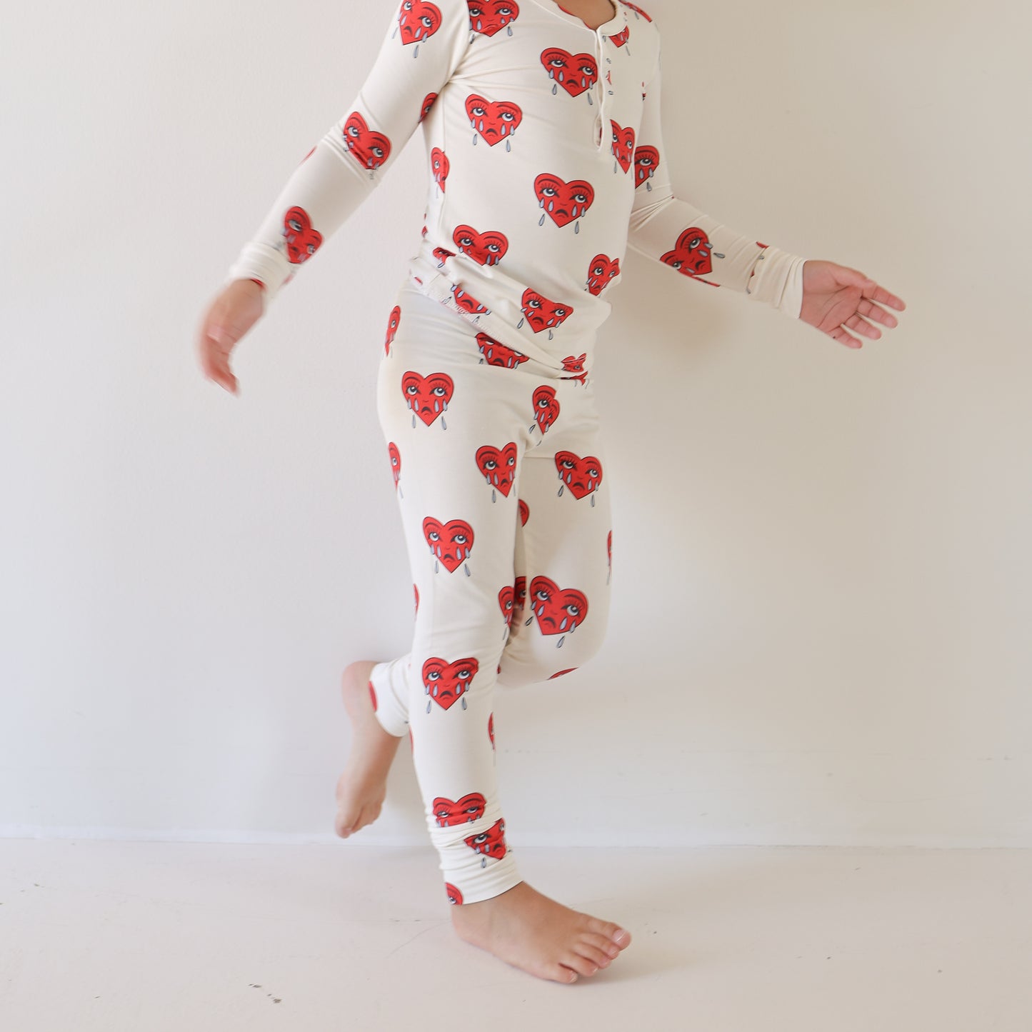 A child stands barefoot against a plain white wall wearing forever french baby’s Bamboo Two Piece Pajamas | Sad Girl—breathable, long-sleeved white sleepwear with red heart-shaped faces. The child's face is not visible in the image.