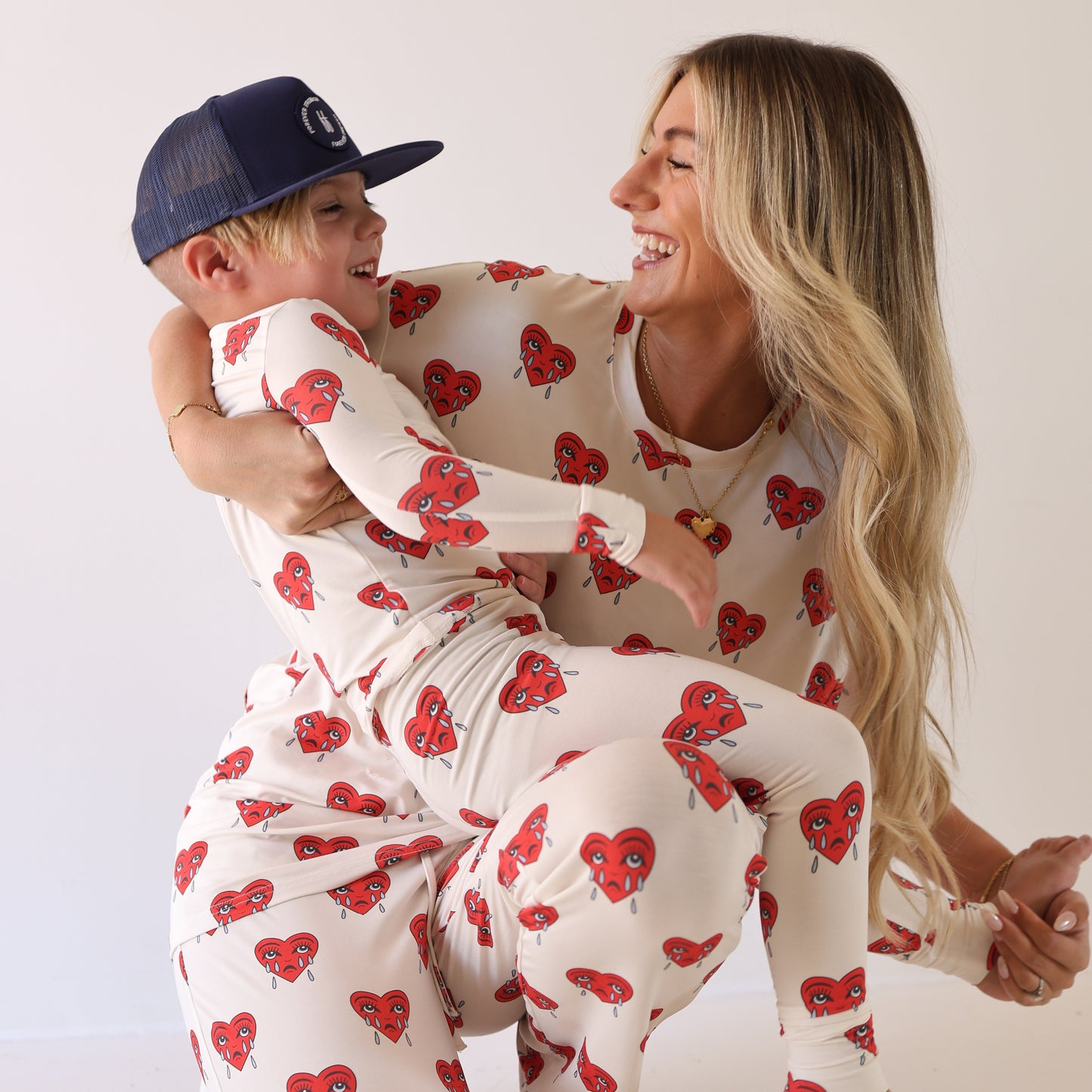 A woman and child laugh together in matching forever french baby “Sad Girl” Short Sleeve Women's Bamboo Pajamas with red heart prints, while the child’s blue cap adds a cute finishing touch.