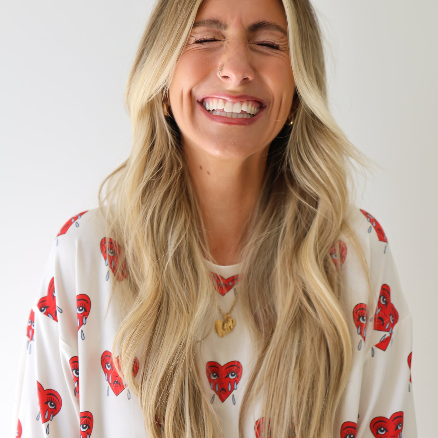A woman with long blonde hair smiles with her eyes closed, wearing the forever french baby Short Sleeve Women's Bamboo Pajamas | Sad Girl—an oversized set featuring red cartoon hearts. She stands against a plain white background.
