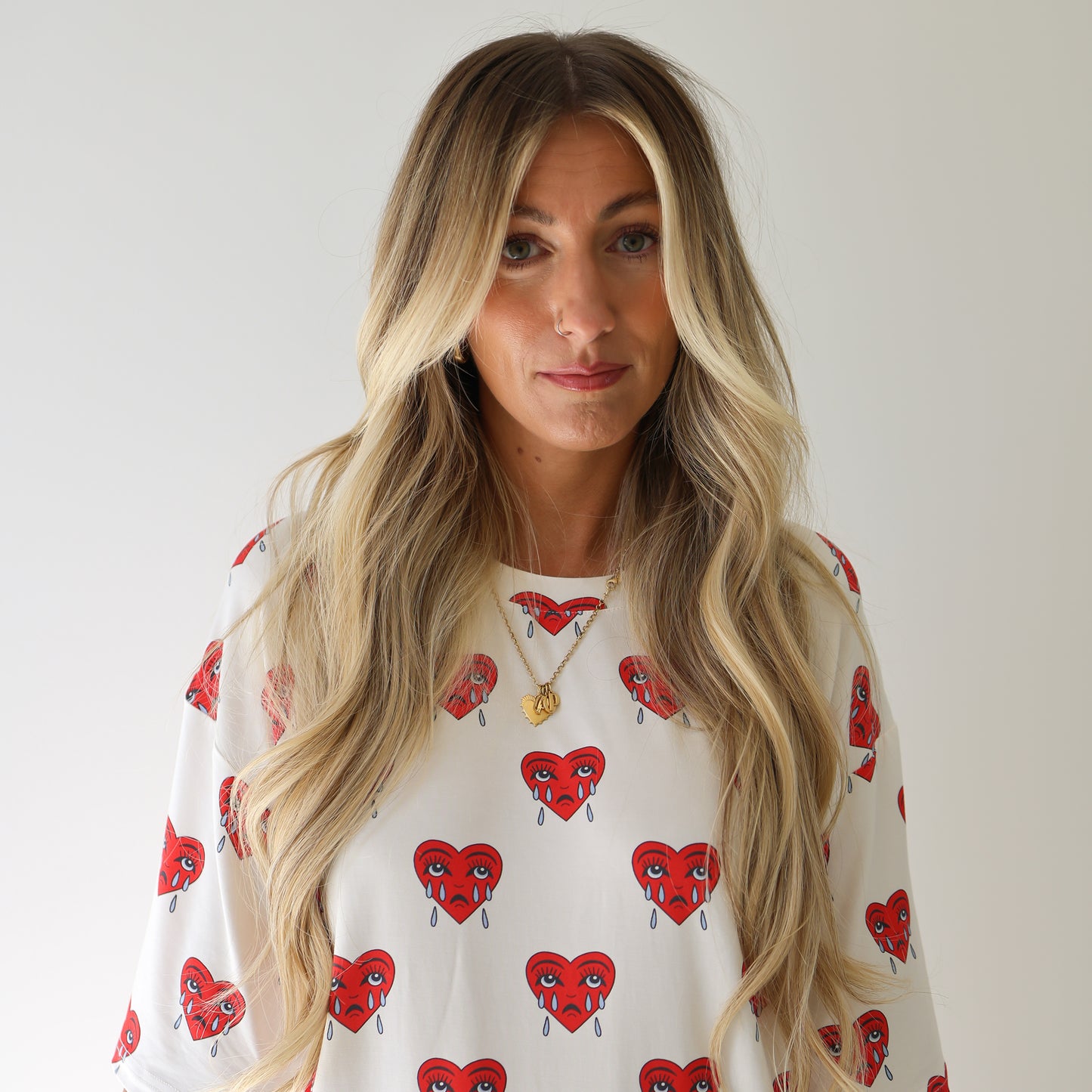 A woman with long, wavy blonde hair and gold necklaces wears the forever french baby Short Sleeve Women’s Bamboo Pajamas | Sad Girl—a white top with red crying heart designs—against a plain light background, looking calm.
