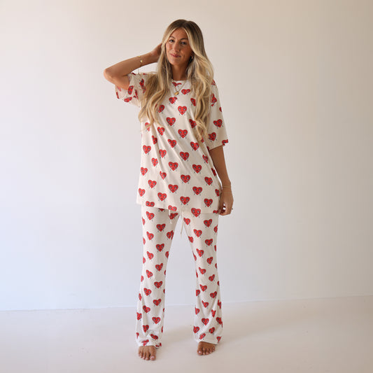 A woman stands barefoot in a plain room, smiling softly at the camera. She wears the forever french baby Short Sleeve Women's Bamboo Pajamas | Sad Girl, featuring red heart patterns, her long blonde hair loose as she touches her head.