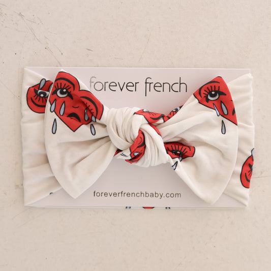 The Bamboo Head Wrap | Sad Girl by forever french baby features a white band with a large bow in the Sad Girl print—red hearts with eyes and teardrops—and comes displayed on a "forever french" card from foreverfrenchbaby.com.