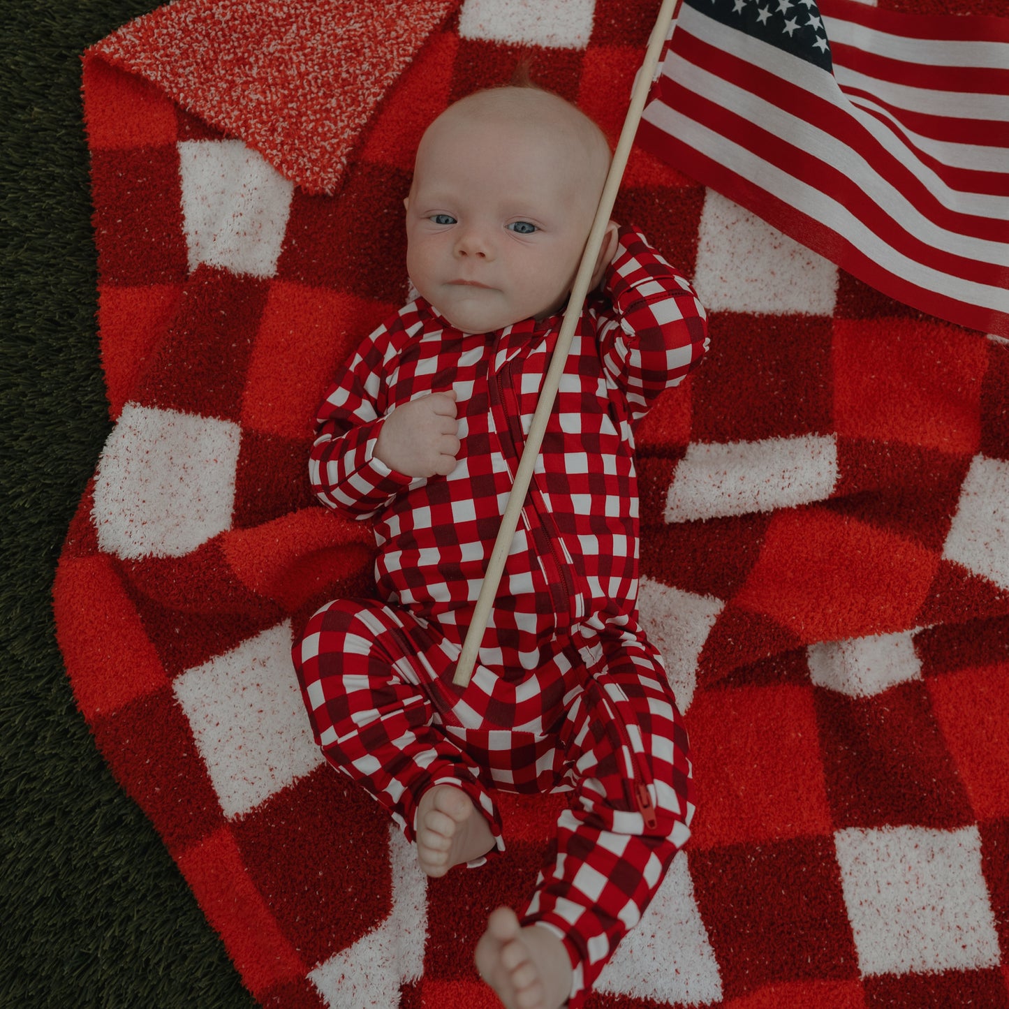 A baby in forever french baby Bamboo Zip Pajamas | Scarlet Gingham lies on a matching blanket, clutching a small American flag, with green grass beside the blanket.