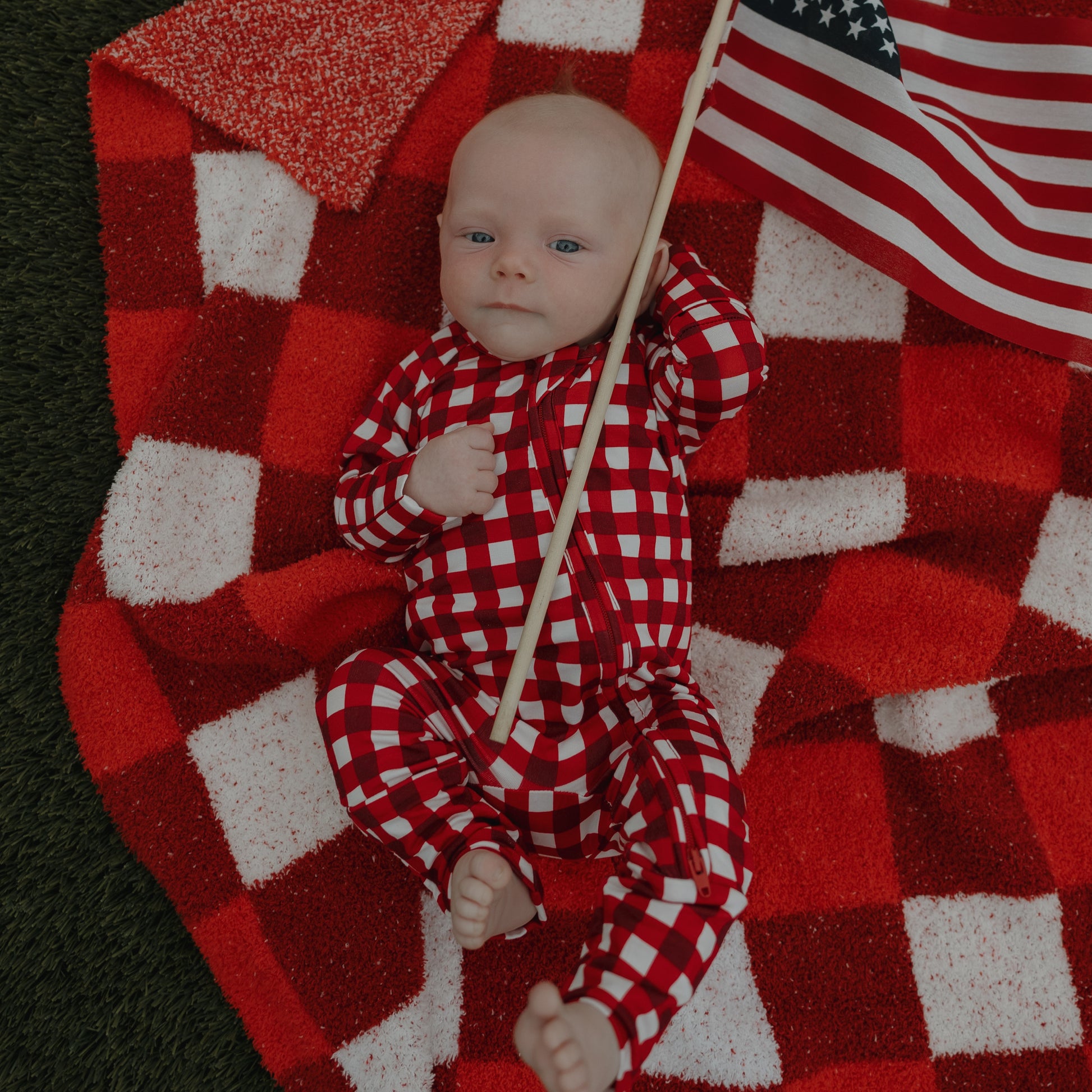 A baby in forever french baby Bamboo Zip Pajamas | Scarlet Gingham lies on a matching blanket, clutching a small American flag, with green grass beside the blanket.