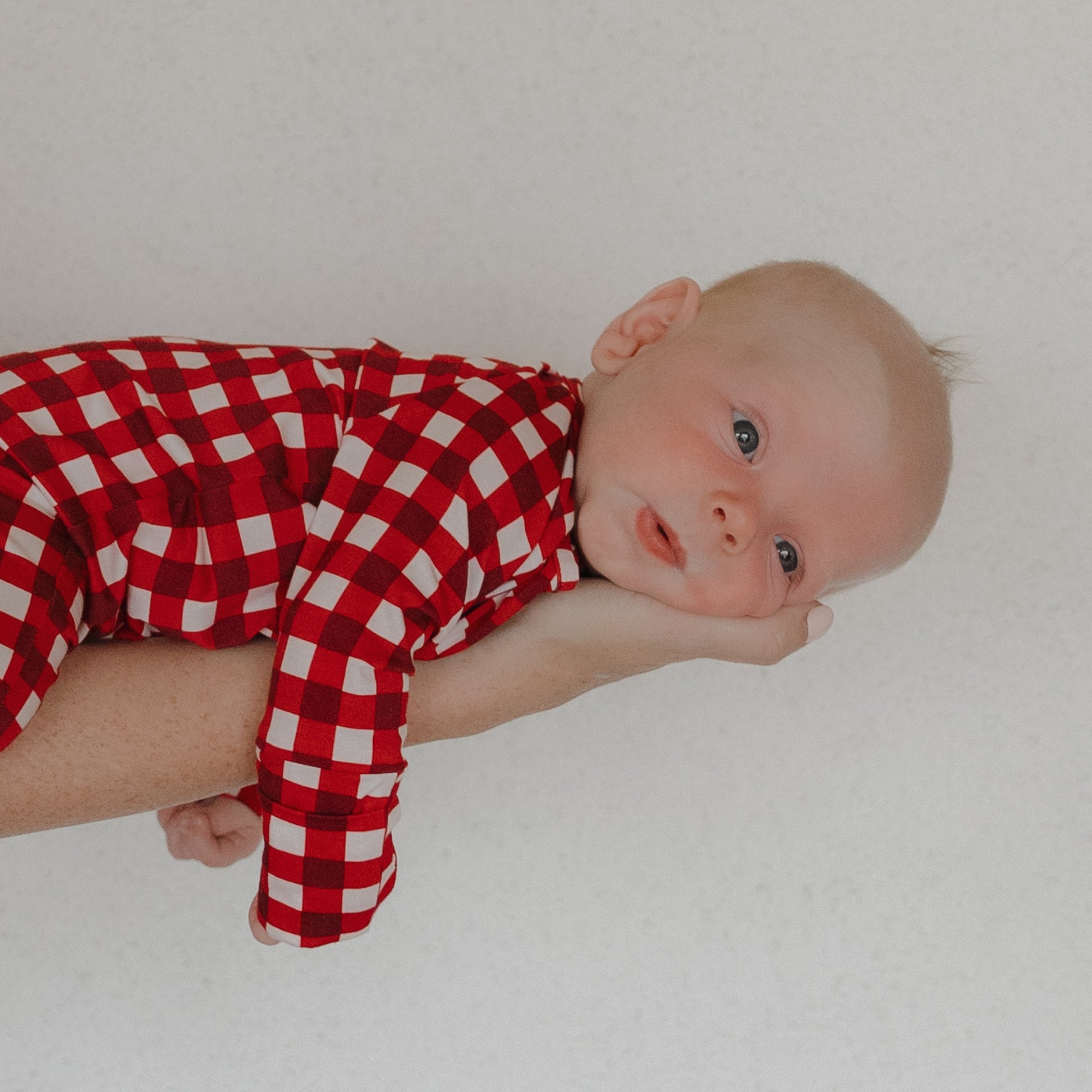 A baby in forever french baby's Bamboo Zip Pajamas in Scarlet Gingham lies on their stomach across an adult's arm, with the adult gently supporting the baby's head against a plain light background.