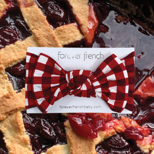 The Bamboo Head Wrap | Scarlet Gingham by forever french baby, featuring a red and white checkered bow, is displayed on a "forever french" card atop a cherry pie with a lattice crust.