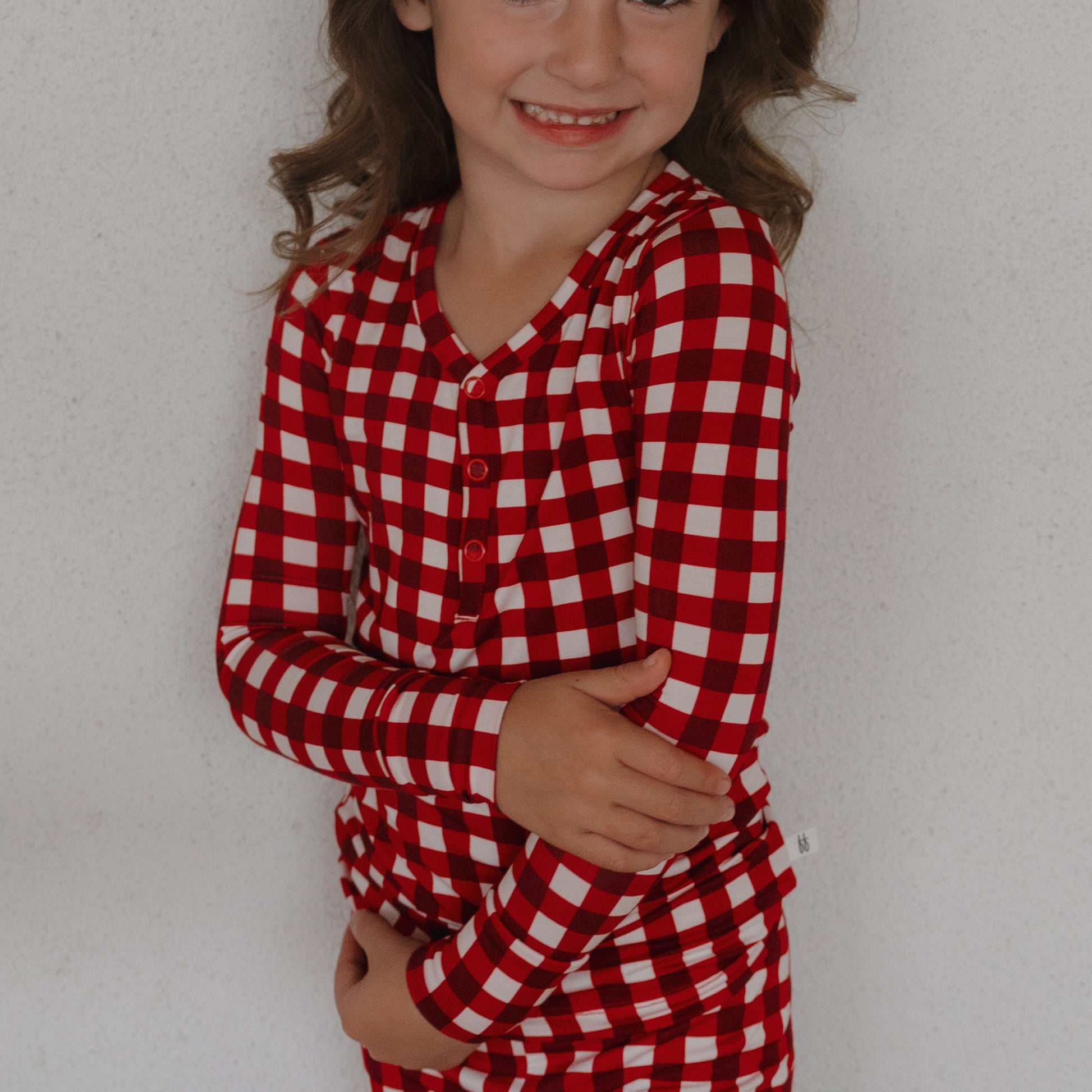 A smiling child with long brown hair wears forever french baby's Bamboo Two Piece Pajamas in Scarlet Gingham, standing against a plain white background with arms gently folded.