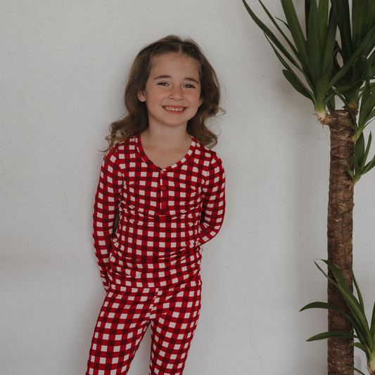 A young girl with long brown hair smiles against a white wall, wearing forever french baby’s Bamboo Two Piece Pajamas in Scarlet Gingham. This hypo-allergenic kids sleepwear is perfect for comfort and style, and she stands beside a tall green indoor plant.