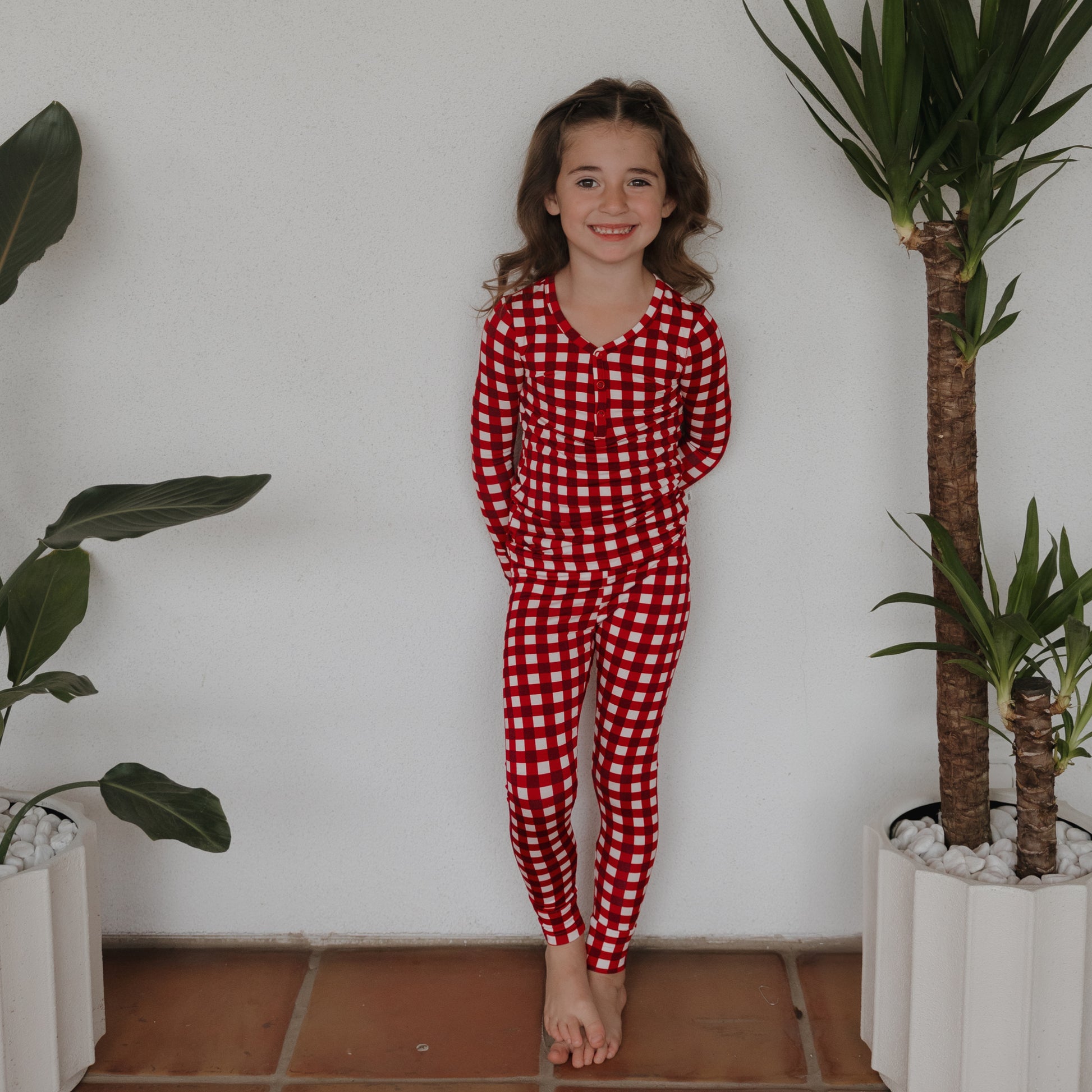 A young girl with long brown hair stands barefoot between two potted plants, smiling in forever french baby's Bamboo Two Piece Pajamas | Scarlet Gingham, in front of a plain white wall.
