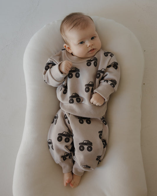 A baby relaxes on a soft white cushion, wearing the forever french baby Child Sweat Set | Monster Truck—an everyday cozy outfit featuring a playful black monster truck print.