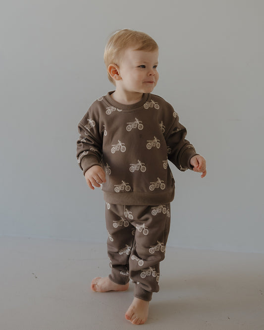A toddler with short blonde hair stands barefoot, wearing the forever french baby Child Sweat Set | Dirt Bike, featuring a fun dirt bike print, against a plain light gray background.