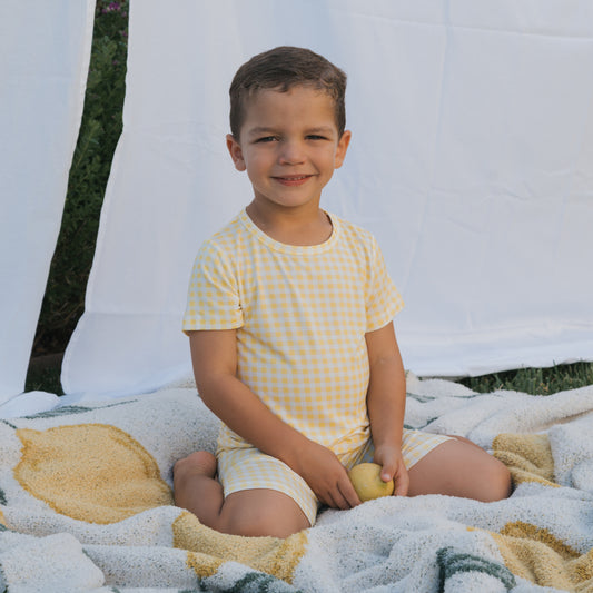 A young boy sits cross-legged on a patterned blanket outdoors, smiling as he holds a yellow fruit. He's wearing the Bamboo Short Set in Citron by forever french baby, with white sheets hanging in the background.