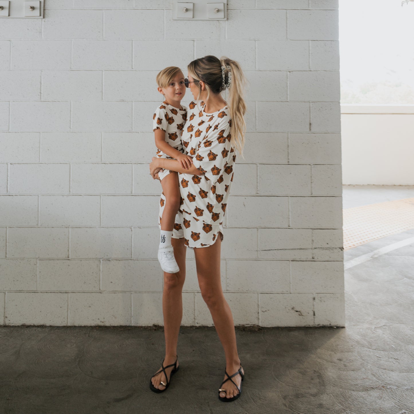 A woman holds a young child, both wearing forever french baby's Bamboo Short Set | Tigers—matching white shirts with brown tiger prints—standing in front of a white brick wall in a bright indoor space.