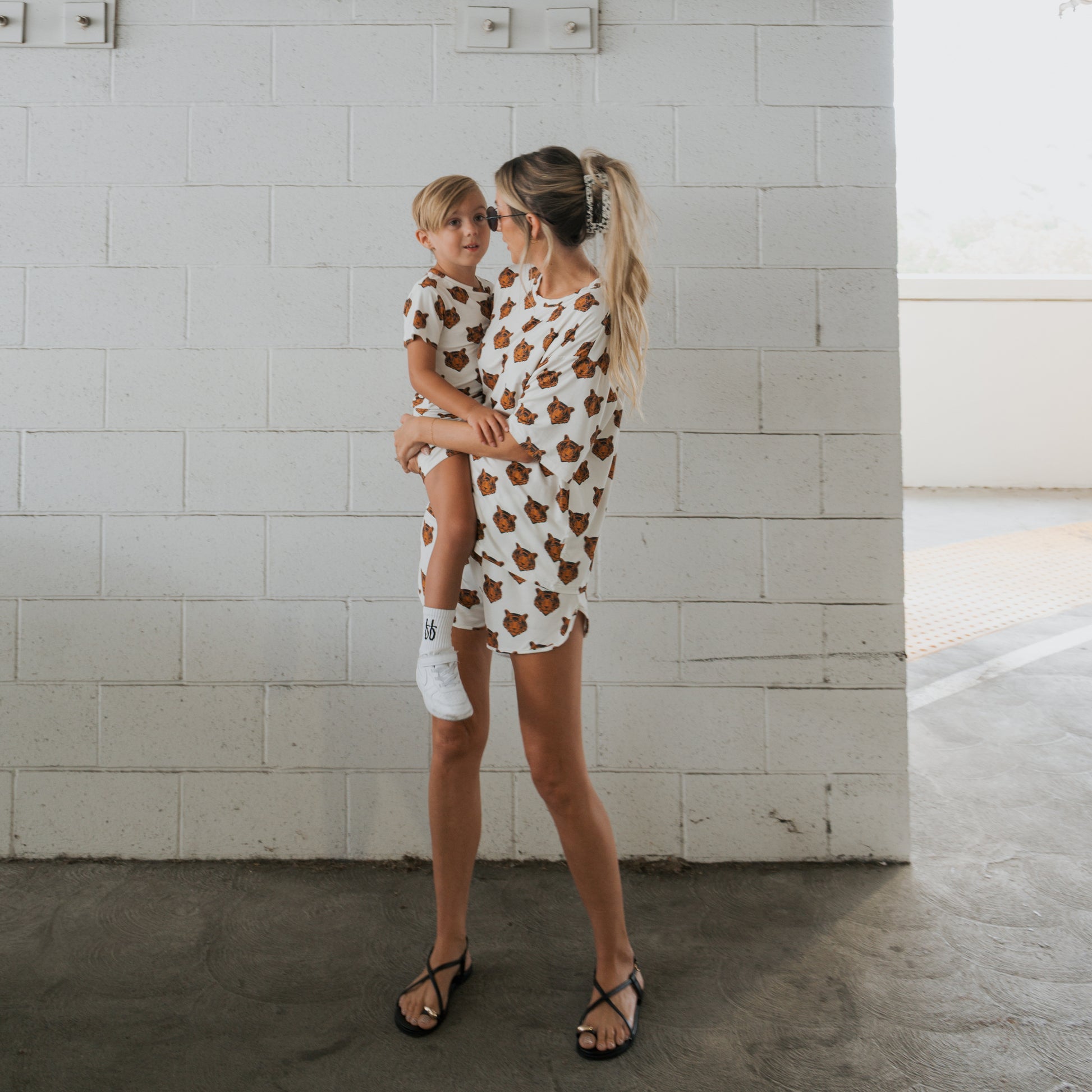 A woman holds a young child, both wearing forever french baby's Bamboo Short Set | Tigers—matching white shirts with brown tiger prints—standing in front of a white brick wall in a bright indoor space.