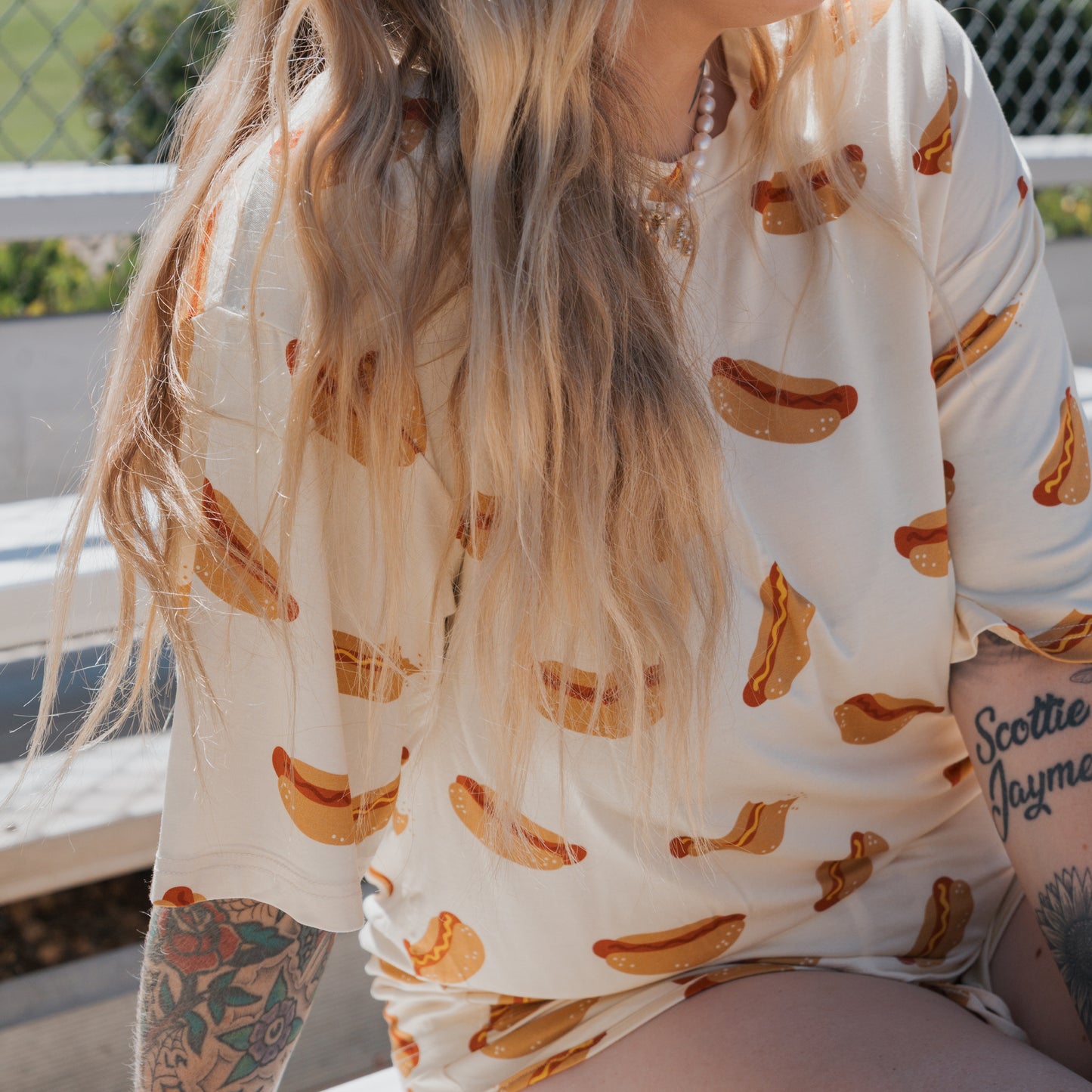 A person with long blonde hair and arm tattoos sits outside on bleachers wearing the forever french baby Women's Bamboo Short Set in the Hot Dogs print.