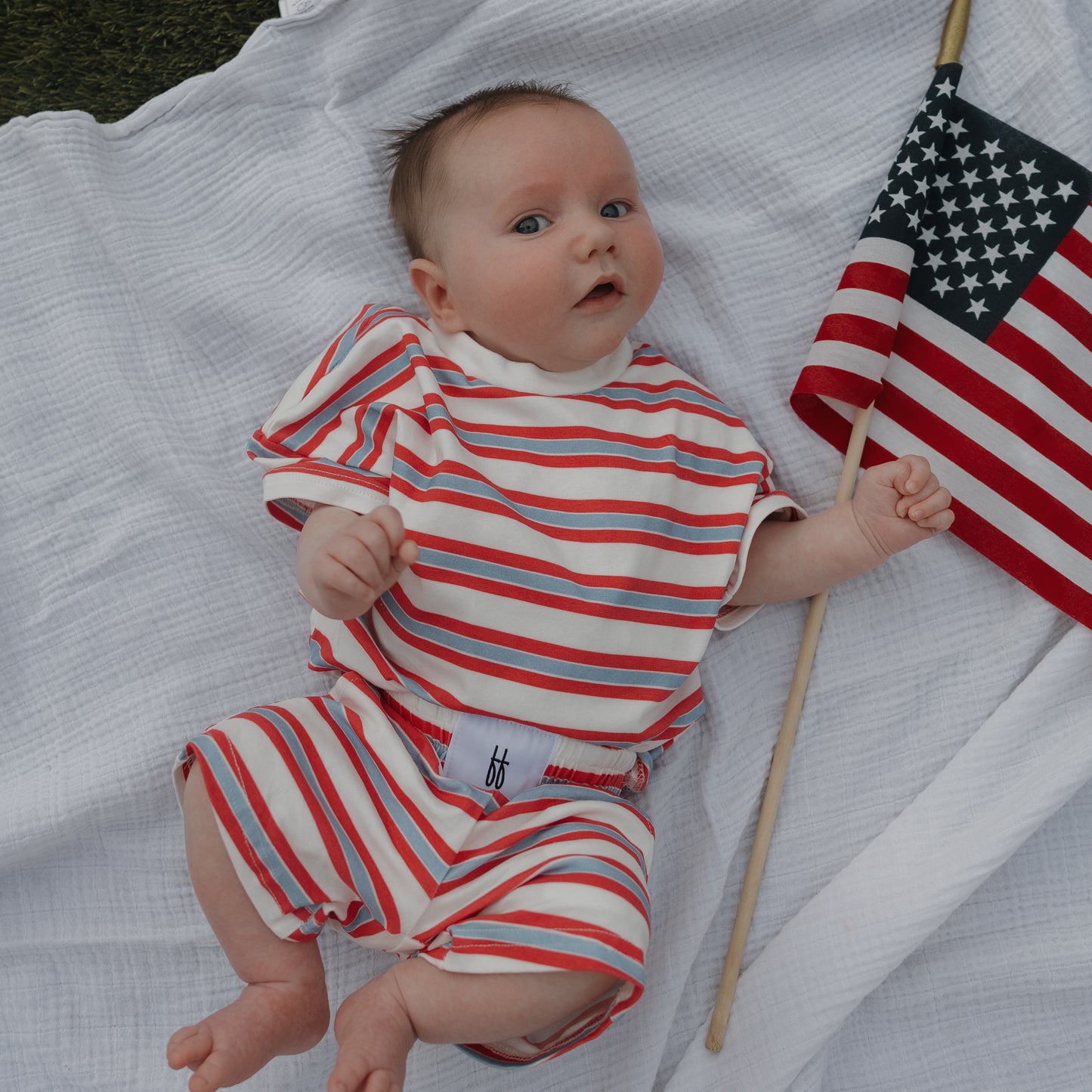 A baby wearing the forever french baby Child Short Set | Heritage Stripe in red, white, and blue lies on a white blanket, holding a small American flag.