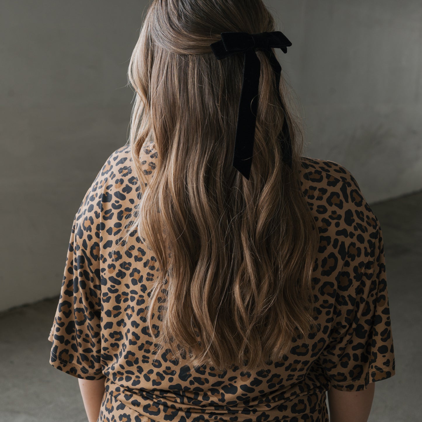 A person with long, wavy light brown hair, wearing a black velvet bow and the Women's Bamboo Short Set | Leopard by forever french baby, stands indoors against a softly lit neutral background.
