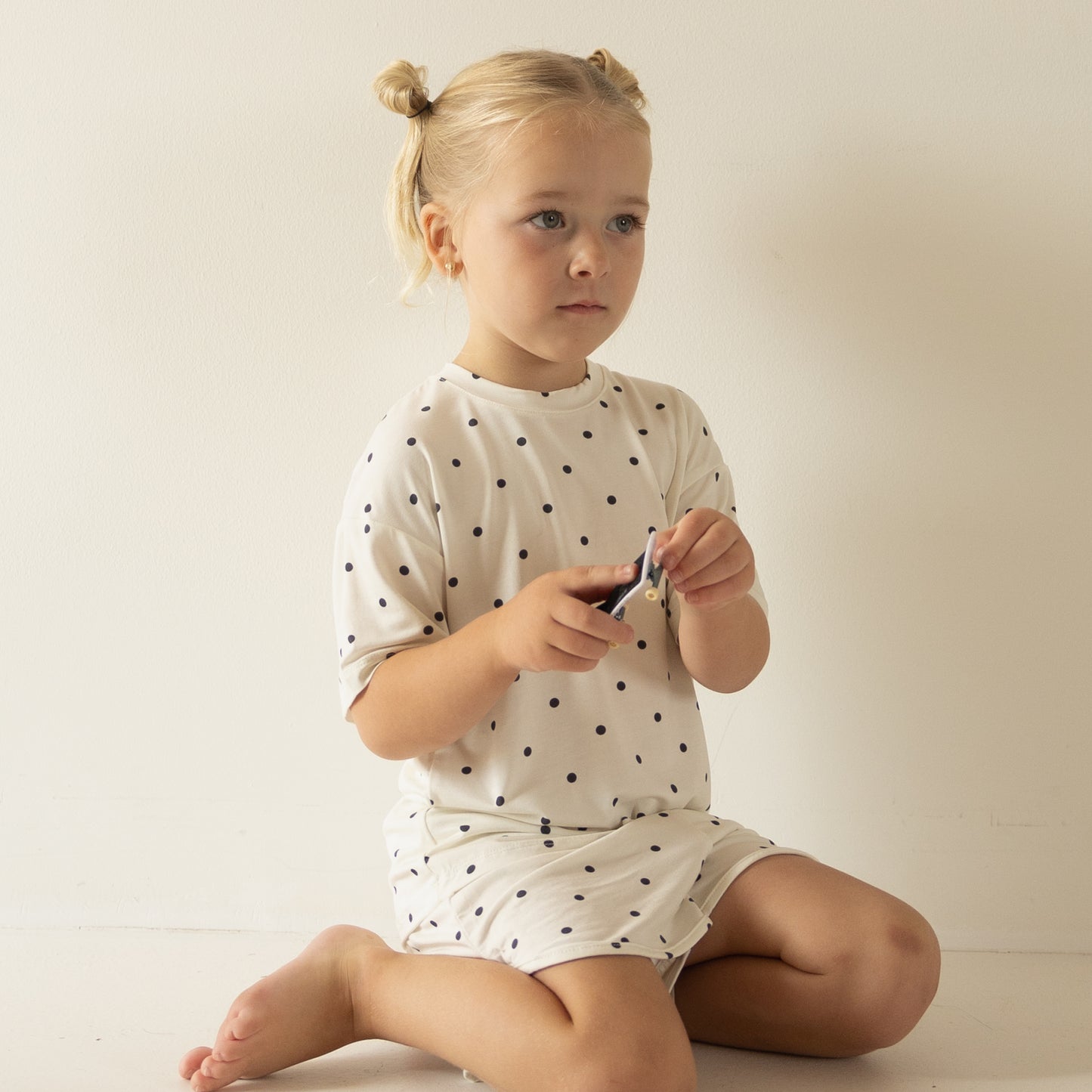 A young blonde girl sits barefoot on the floor, wearing the forever french baby Girl's Bamboo Short Set | Annie, a white polka-dot, breathable outfit, looking slightly to the side and holding a small object.
