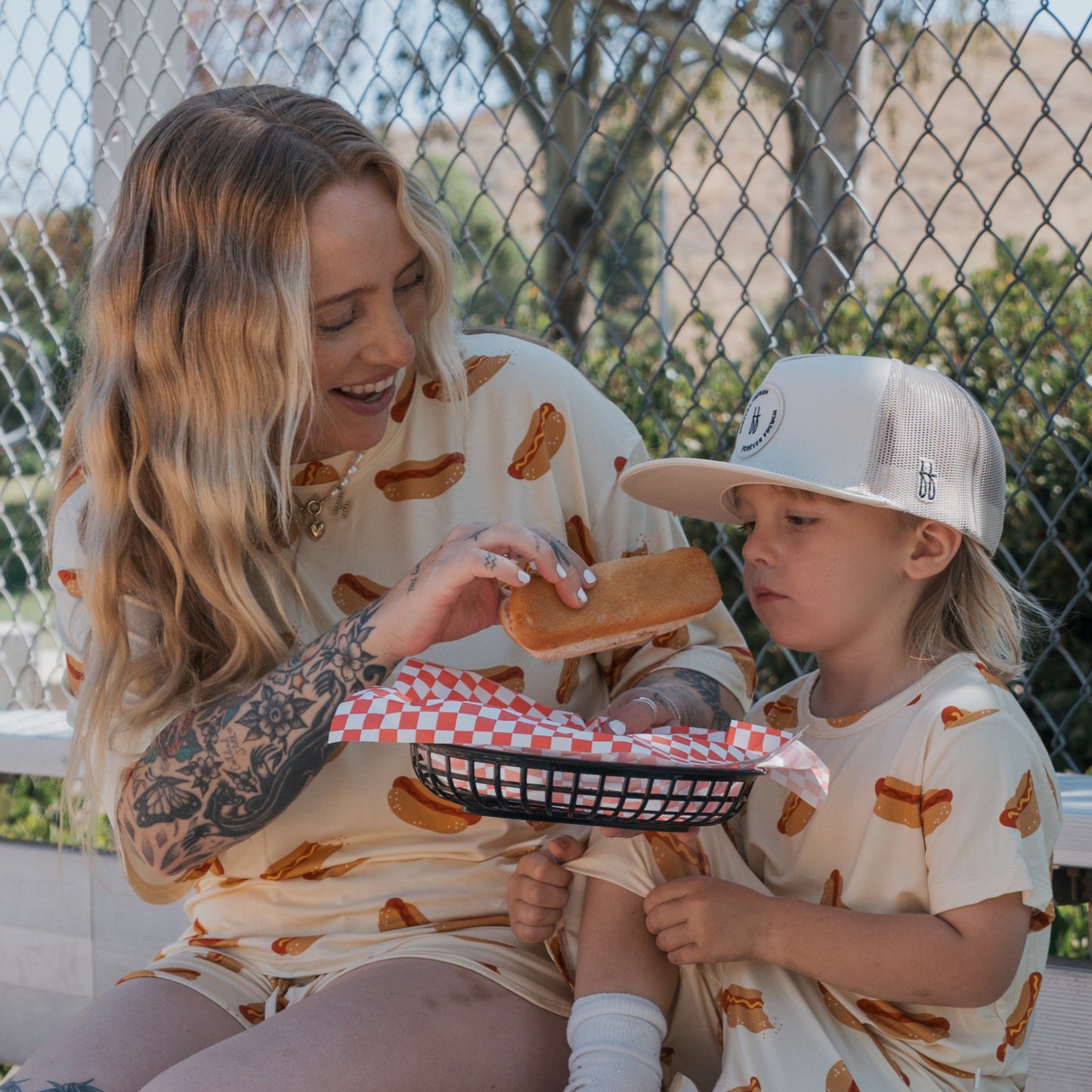 A smiling woman and young boy sit outdoors in forever french baby's Women's Bamboo Short Set | Hot Dogs. The soft, breathable matching outfits are perfect for summer fun as they share a meal together.