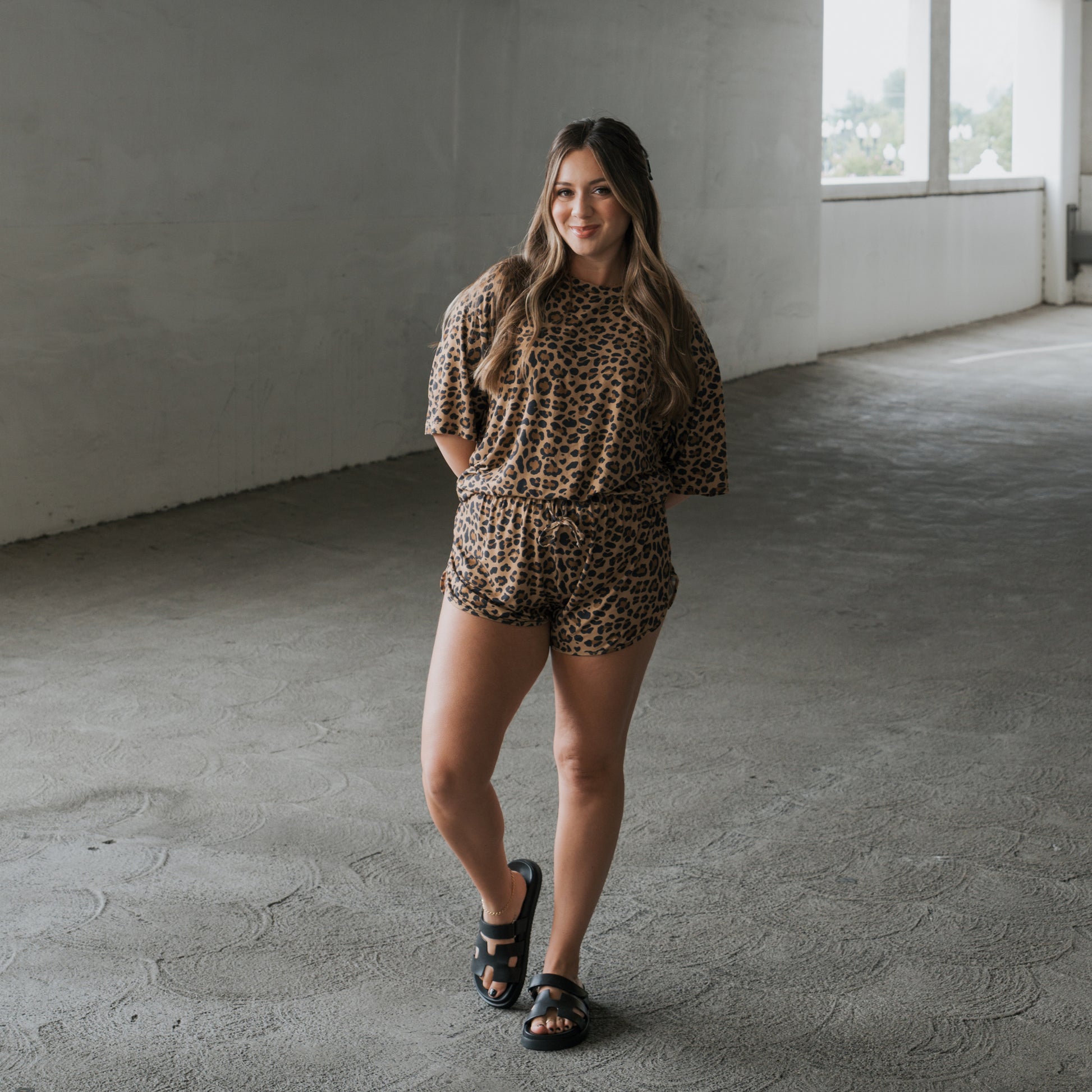A woman with long hair wears the Women's Bamboo Short Set in Leopard by forever french baby and black sandals, standing on a concrete floor in a bright, open indoor space, smiling slightly at the camera.
