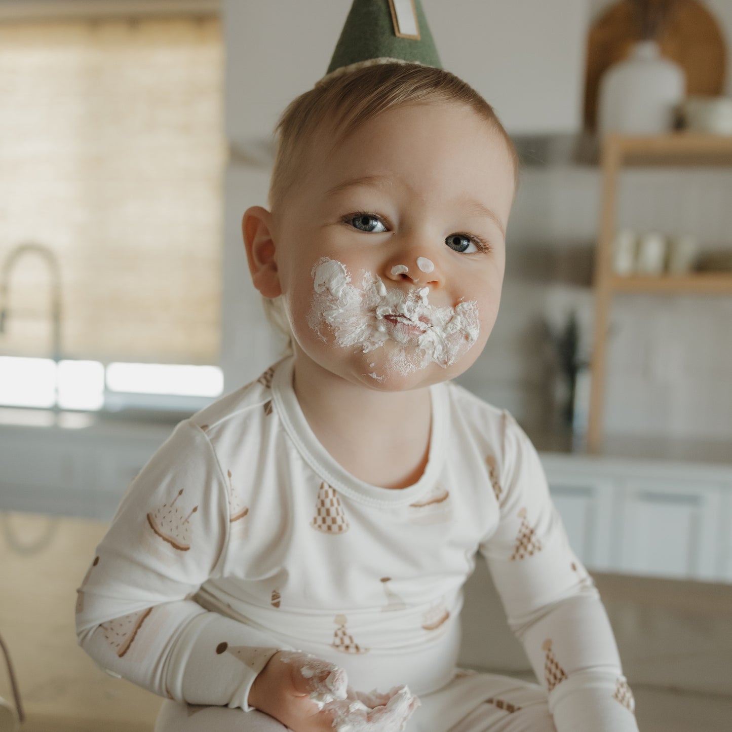 A toddler wearing a party hat sits on a counter with white frosting on their face and hands, looking at the camera in the forever french baby Child Sleepy Time Set | Birthday Boy.