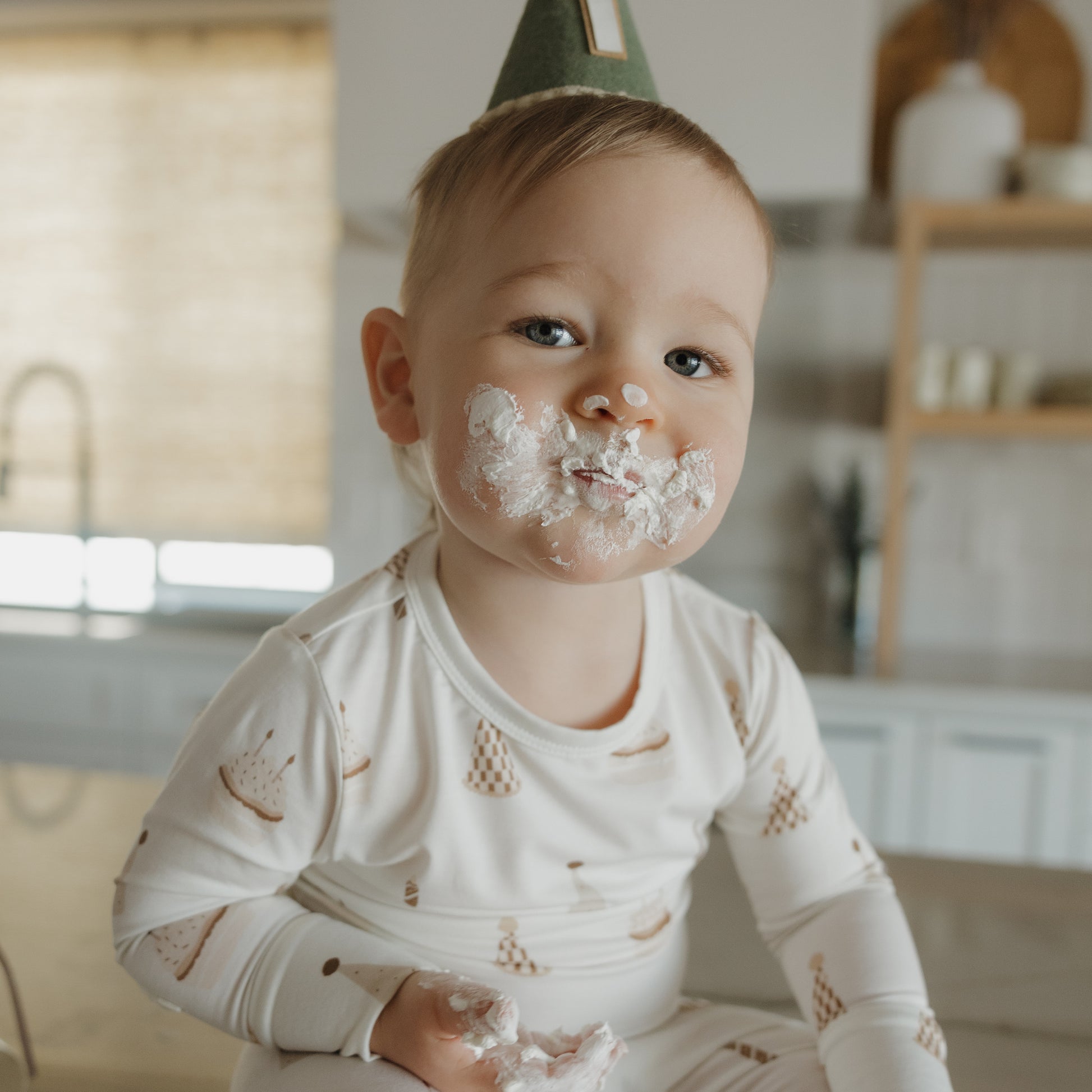 A toddler wearing a party hat sits on a counter with white frosting on their face and hands, looking at the camera in the forever french baby Child Sleepy Time Set | Birthday Boy.