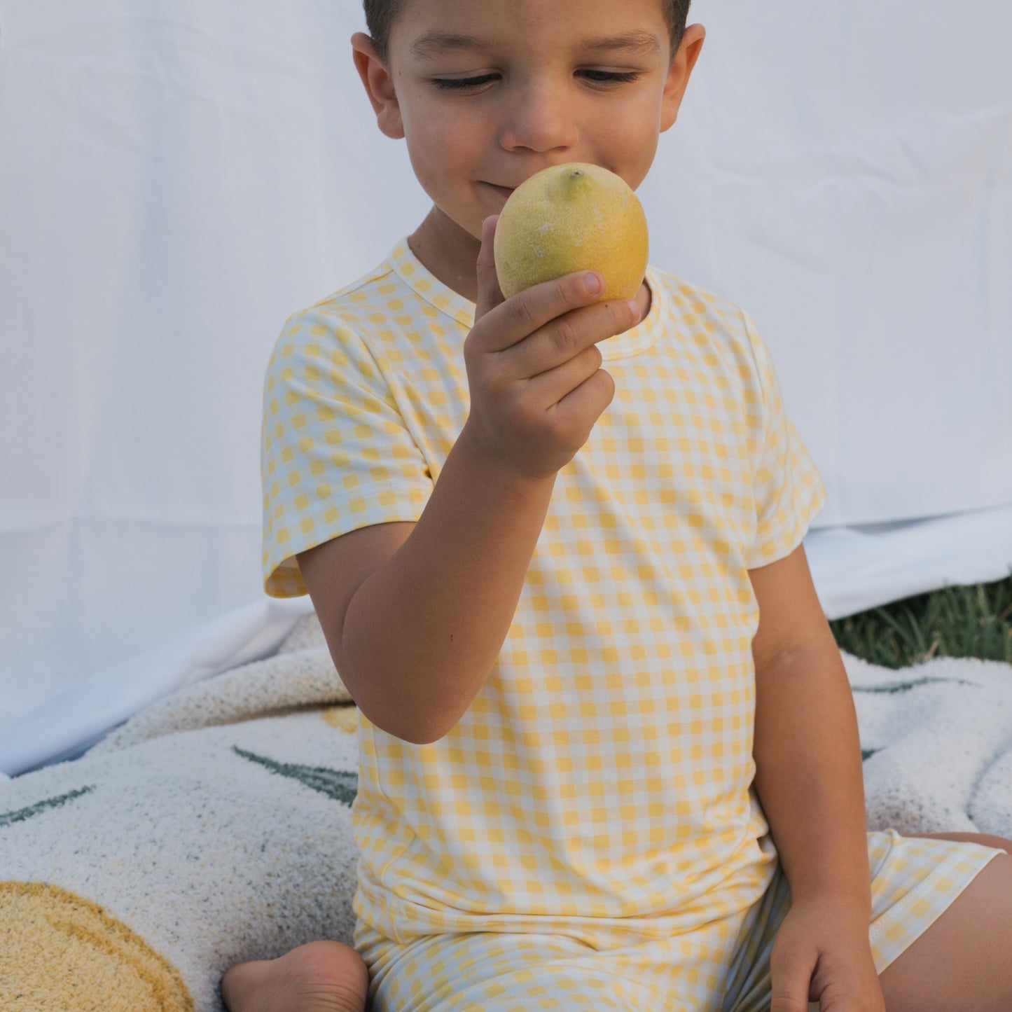 A young child in the forever french baby Bamboo Short Set | Citron sits outdoors on a blanket, holding and smelling a lemon against a white sheet backdrop, wearing yellow and white checkered pajamas.
