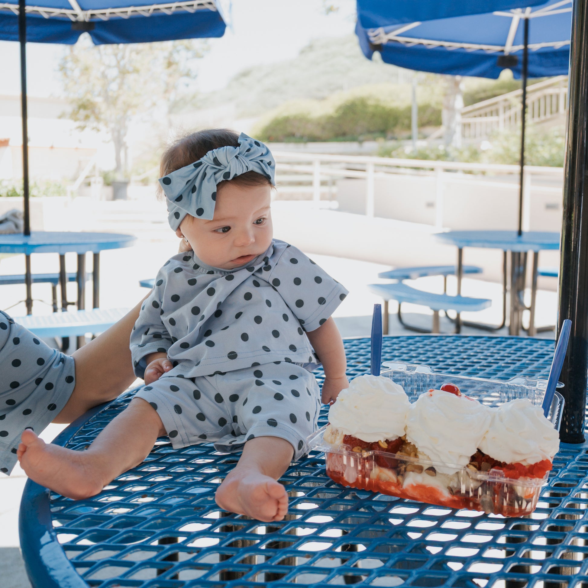 A baby wearing the forever french baby "Child Short Set | Sully" sits on a blue metal table, gazing at a tray of strawberry shortcake with whipped cream on a sunny patio.