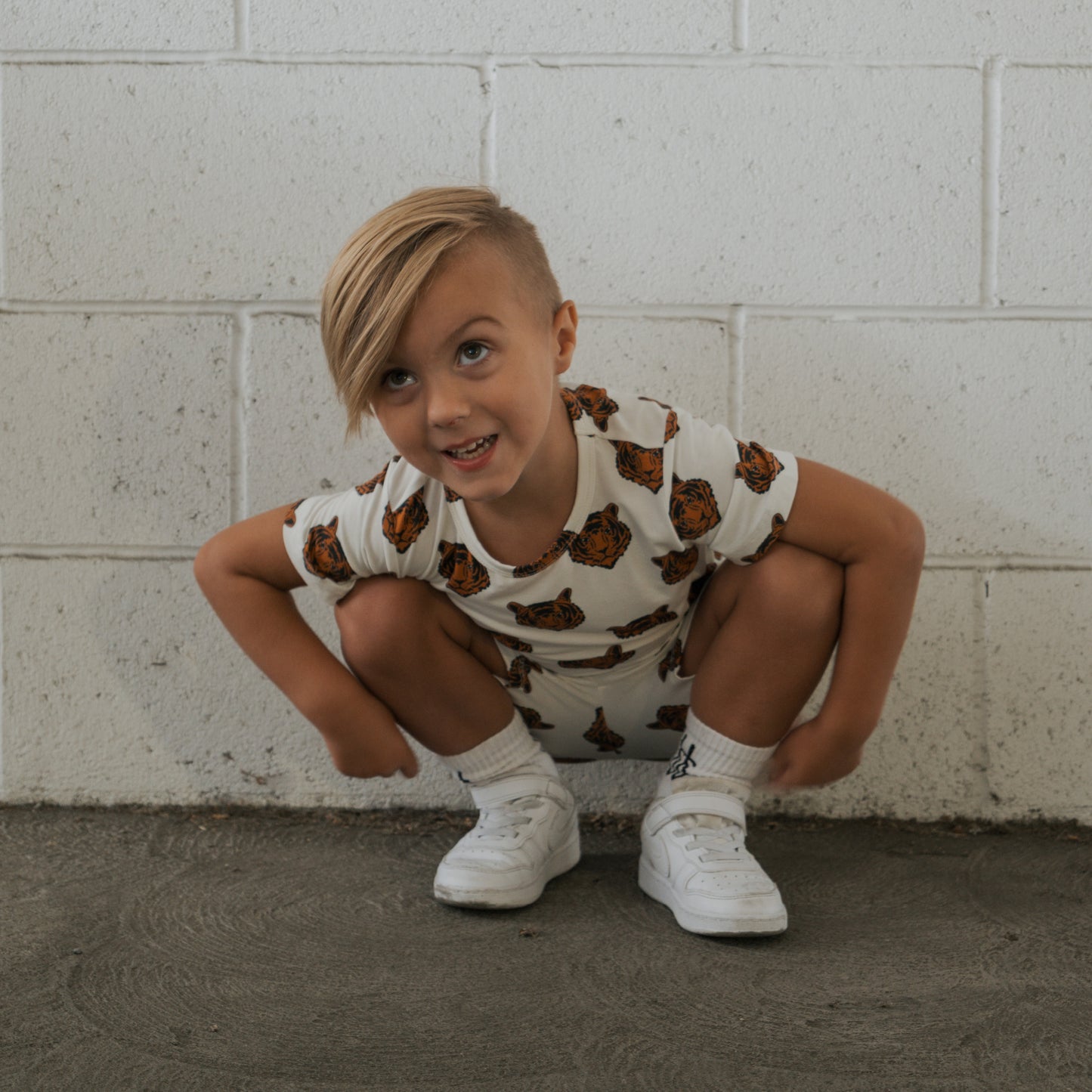 A young child with blonde hair squats in front of a white brick wall, smiling upward while wearing the forever french baby Bamboo Short Set | Tigers—hypoallergenic kids clothing featuring a tiger face print—paired with white socks and sneakers.