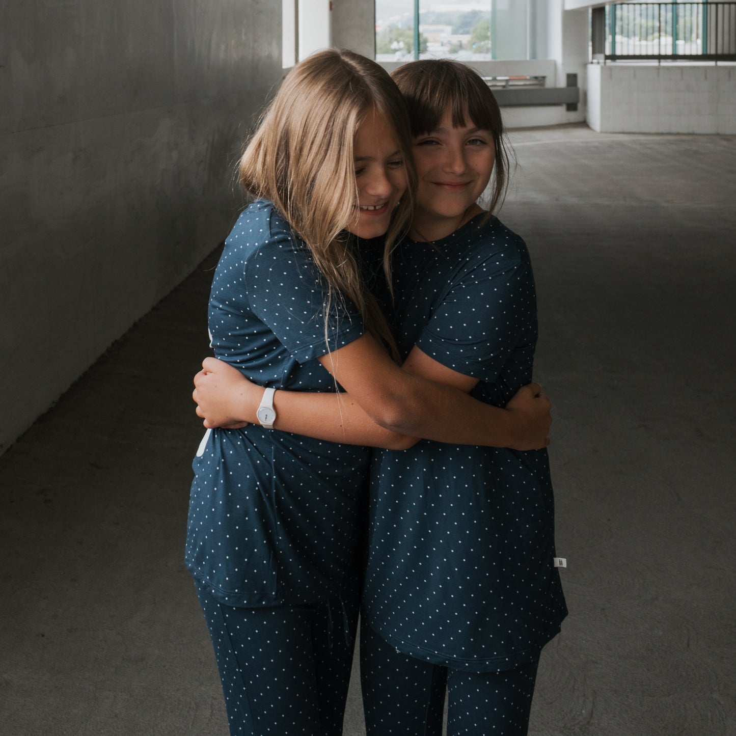 Two young girls wear matching forever french baby Child Short Sleeve Bamboo Lounge Set | Emery in navy polka dot as they hug and smile together in a covered outdoor space with concrete floors and walls.