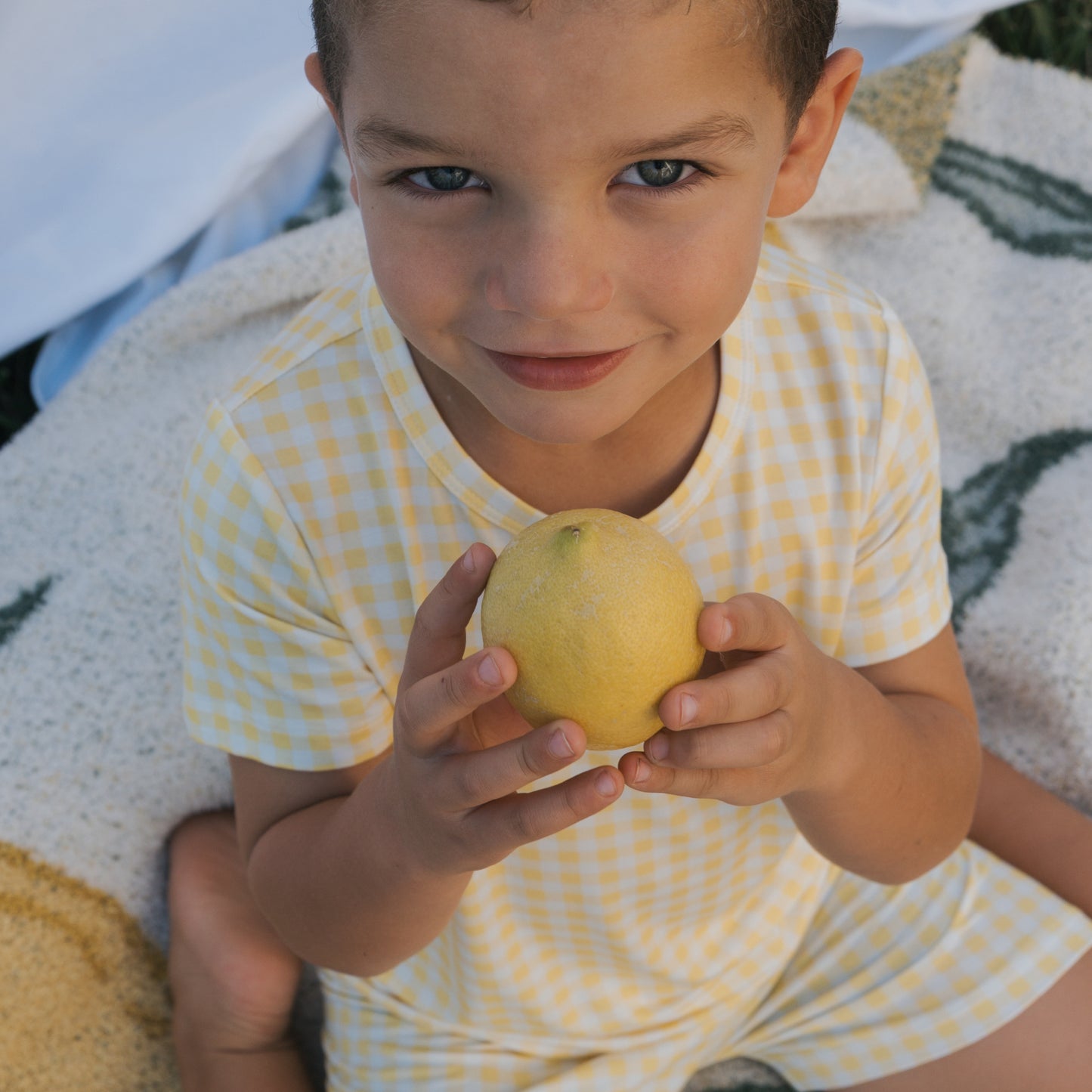 A young child wears the Bamboo Short Set in Citron by forever french baby, sitting on a patterned blanket, holding a lemon with both hands and smiling up at the camera.
