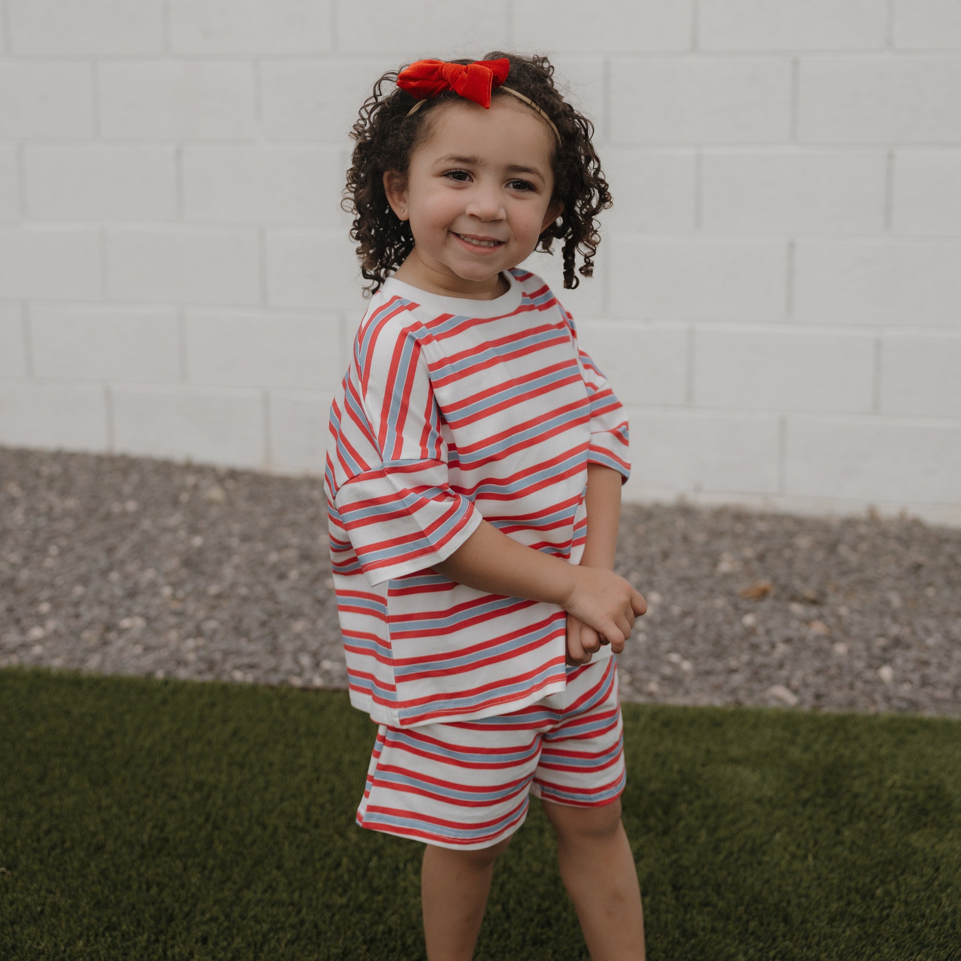A young child with curly hair and a red bow smiles outdoors, wearing the forever french baby Child Short Set | Heritage Stripe, standing on grass in front of a white brick wall.