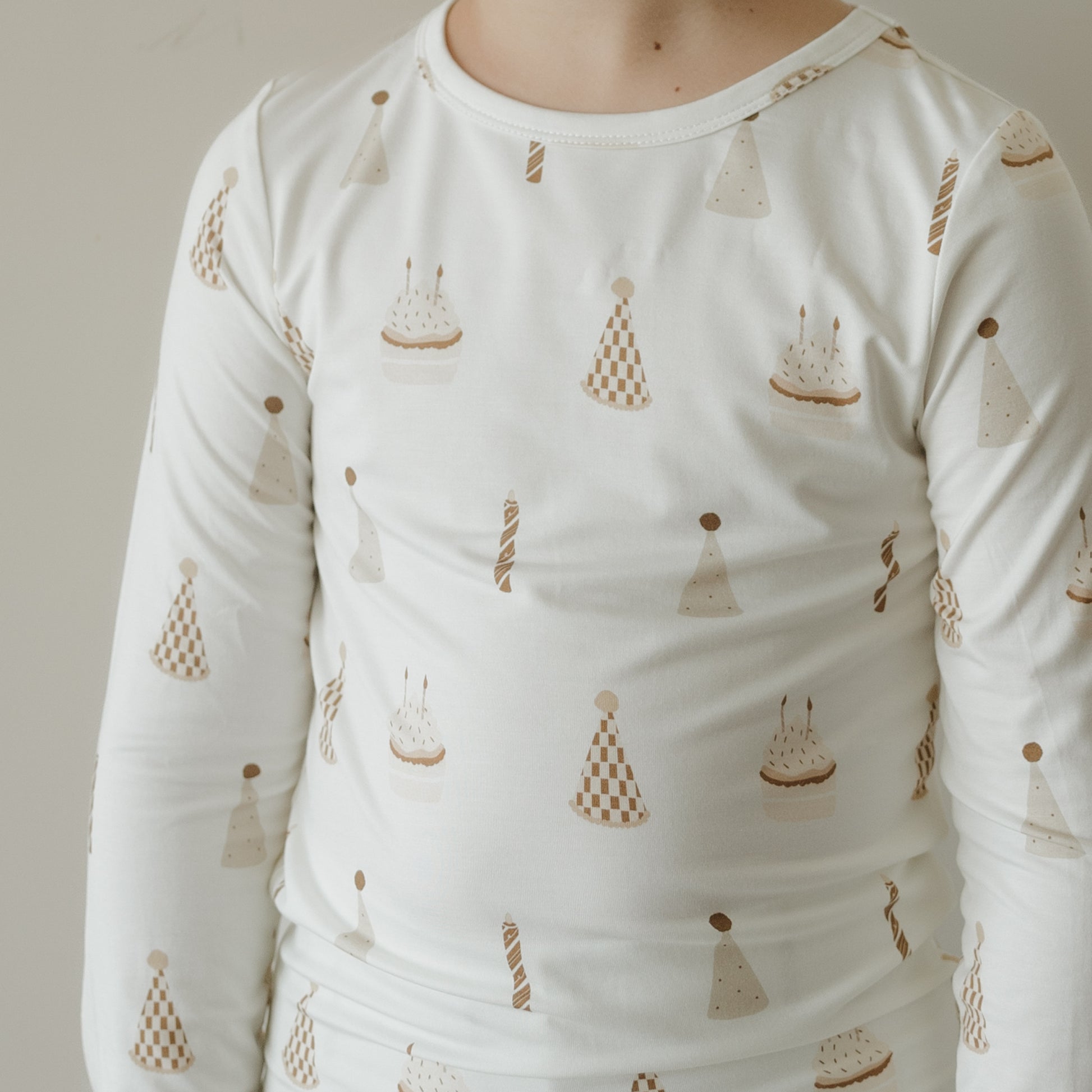 A child stands against a plain light background wearing the forever french baby Child Sleepy Time Set | Birthday Boy—a white long-sleeve shirt adorned with beige party hats, cupcakes, and candles—showcasing hypo-allergenic children's clothing that is gentle on sensitive skin.