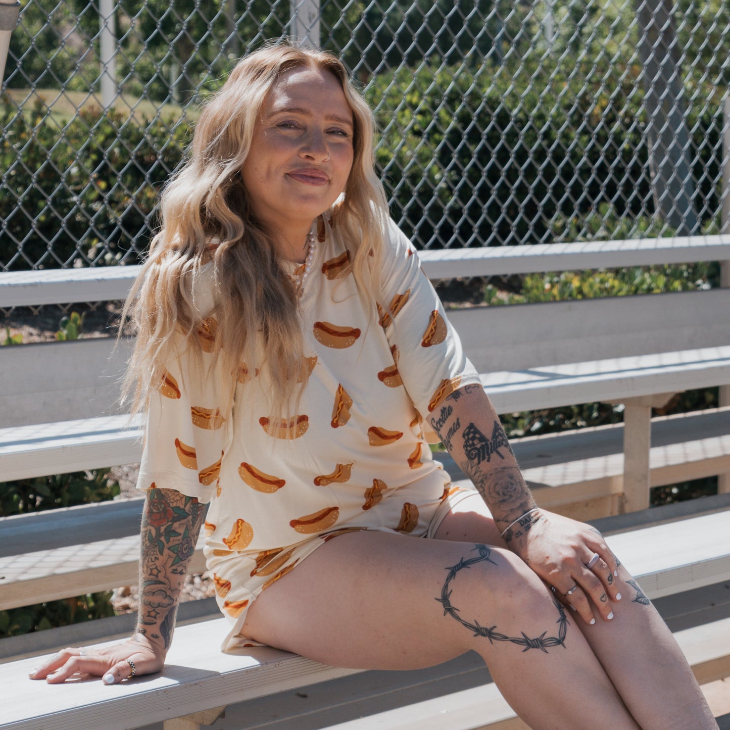 A woman with long, wavy blonde hair sits on bleachers, smiling and showing tattoos. She wears the forever french baby Women's Bamboo Short Set | Hot Dogs. A chain-link fence and greenery are in the background.