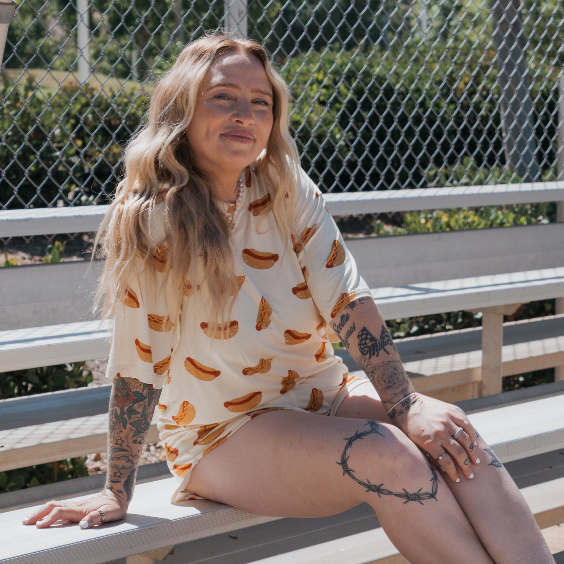 A woman with long, wavy blonde hair sits on bleachers, smiling and showing tattoos. She wears the forever french baby Women's Bamboo Short Set | Hot Dogs. A chain-link fence and greenery are in the background.