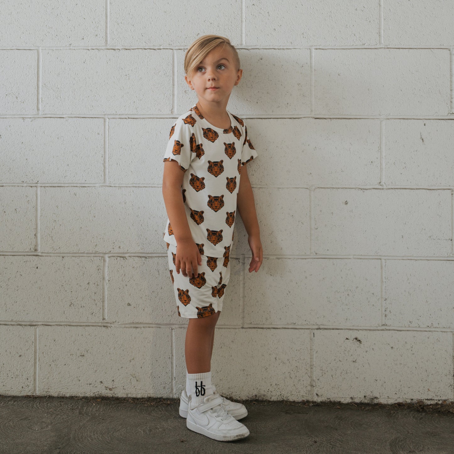 A young blonde child stands against a white brick wall, wearing the forever french baby Bamboo Short Set | Tigers, paired with white socks and sneakers, looking slightly upward to the left.