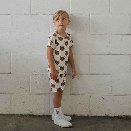 A young blonde child stands against a white brick wall, wearing the forever french baby Bamboo Short Set | Tigers, paired with white socks and sneakers, looking slightly upward to the left.