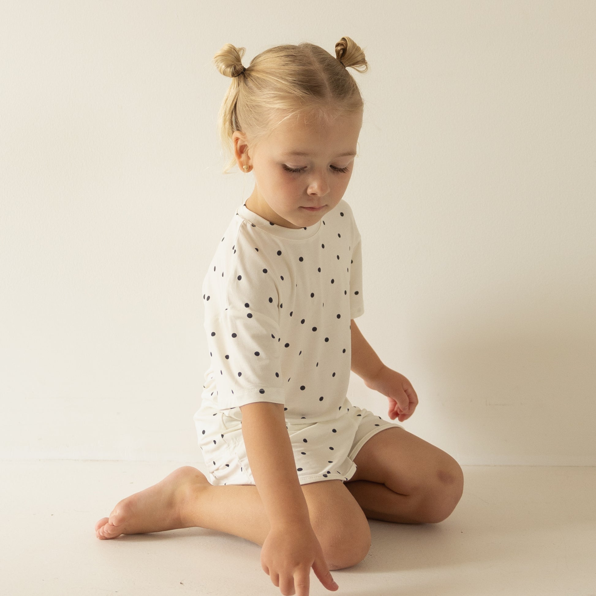 A young girl with blonde hair in two buns wears the forever french baby Girl's Bamboo Short Set | Annie in white with black polka dots, sitting on the floor against a plain background, looking down and touching the ground.